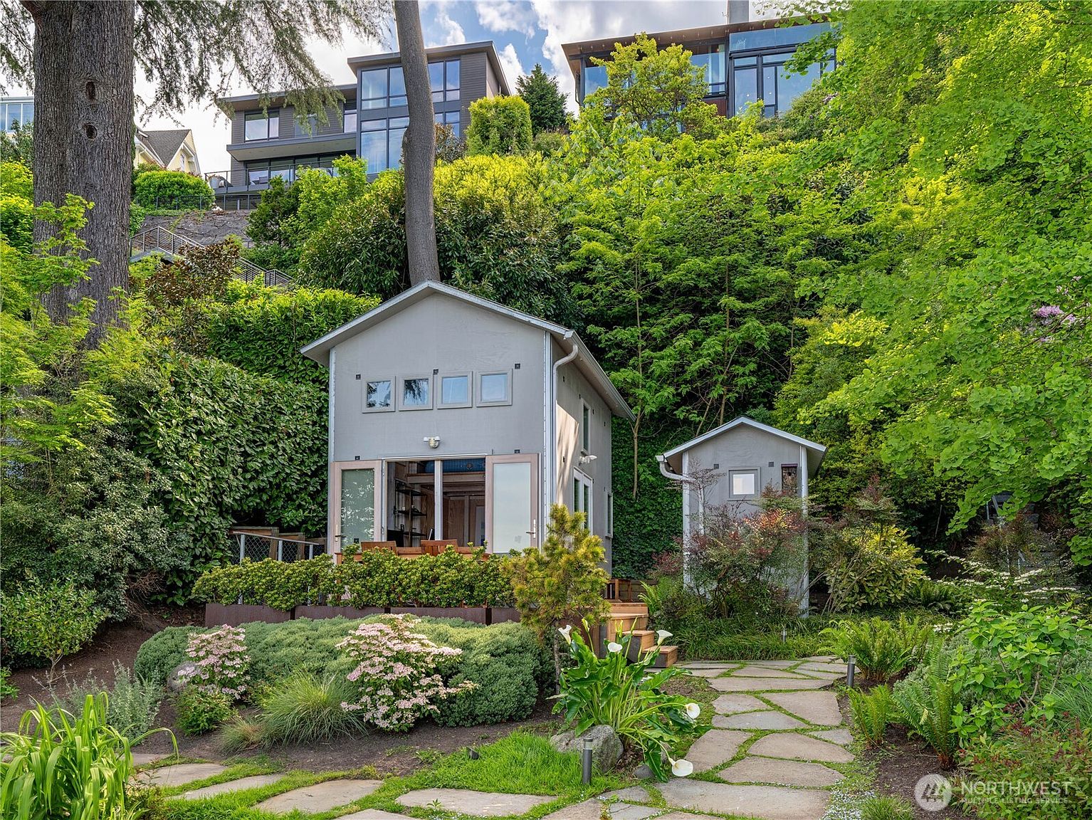 This image captures a serene, tiered garden setting featuring a small, light-gray detached studio or guest cottage nestled into a lush, green hillside. A stone-paved path leads toward the structure, which is surrounded by vibrant landscaping, including flowering shrubs and mature trees. In the background, a larger, modern multi-story residence with expansive glass windows sits perched at the top of the slope, creating a dramatic contrast between the intimate garden retreat and the elevated main home.