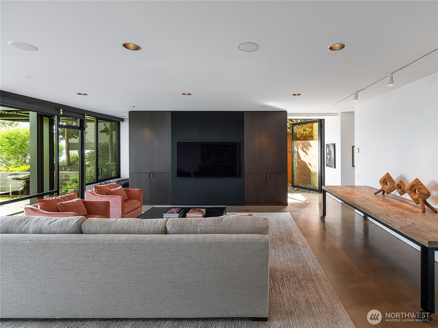 This modern living room features a sleek, dark-toned media wall with a built-in television and integrated cabinetry. A large, light-colored sofa and two coral-toned armchairs are arranged on a textured area rug, while a long, rustic wooden console table sits to the right. The space is illuminated by recessed lighting and benefits from expansive windows that provide a view of the outdoor greenery.