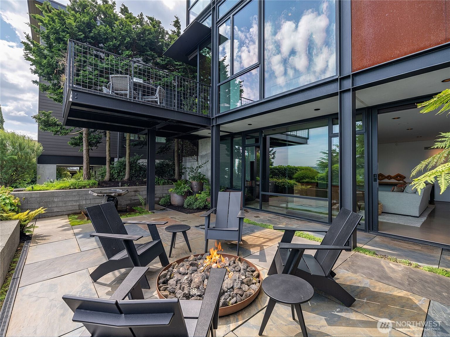 This modern outdoor patio features a circular fire pit surrounded by four dark-toned Adirondack chairs and small side tables, set upon a stone-tiled surface. The space is framed by a striking industrial-style structure with large glass panels and a cantilevered balcony above, creating a sophisticated indoor-outdoor living experience. Lush greenery and mature trees in the background provide a private, serene atmosphere for this contemporary home.