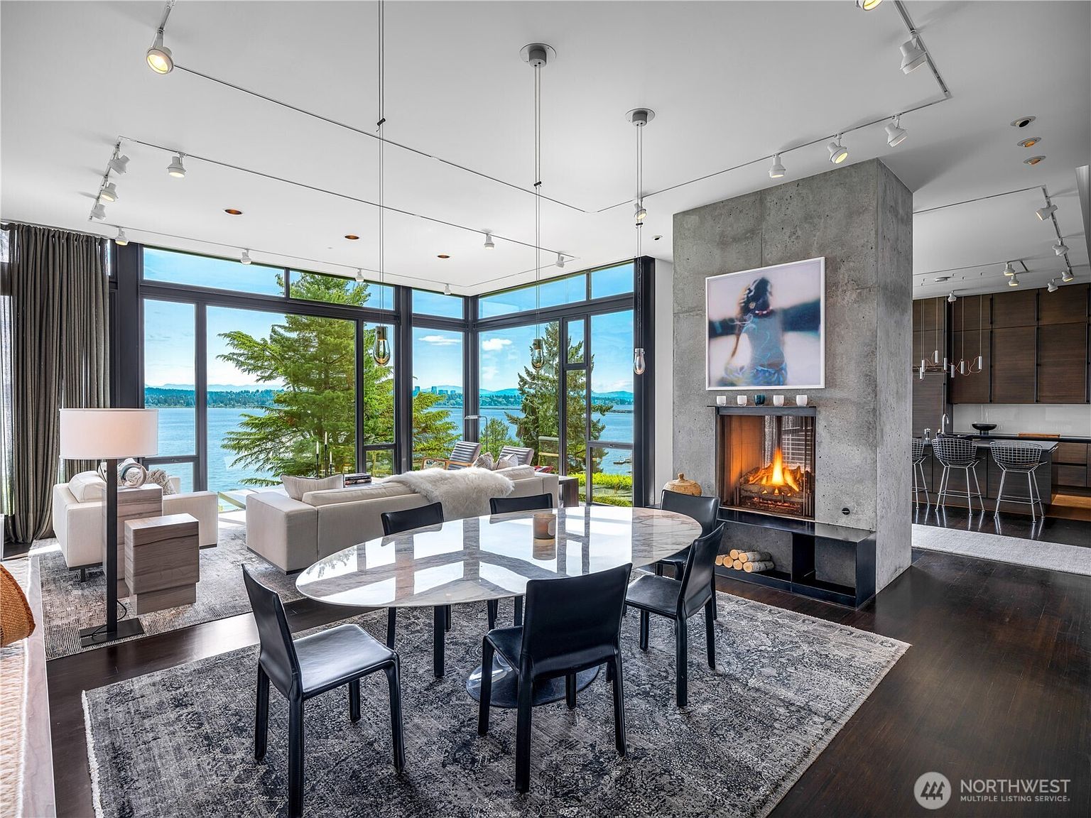This open-concept living and dining area features a sophisticated, modern aesthetic with dark wood flooring and a prominent concrete-clad chimney breast. A sleek oval dining table sits on a patterned rug, while large windows provide expansive, unobstructed views of the water and surrounding trees. The space is illuminated by a combination of track lighting and minimalist pendant fixtures, creating a bright and airy atmosphere.