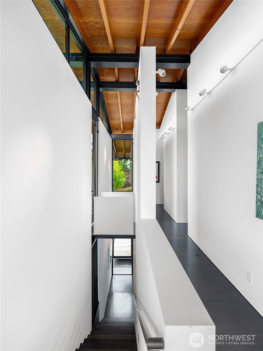 This striking interior view captures a modern staircase and hallway characterized by high-contrast design and architectural depth. The space features a dark, sleek staircase leading downward, flanked by crisp white walls and exposed wooden ceiling beams supported by black steel structural elements. A narrow, elevated walkway with a solid white railing overlooks the stairwell, creating a dramatic, multi-level perspective that emphasizes clean lines and contemporary aesthetics.
