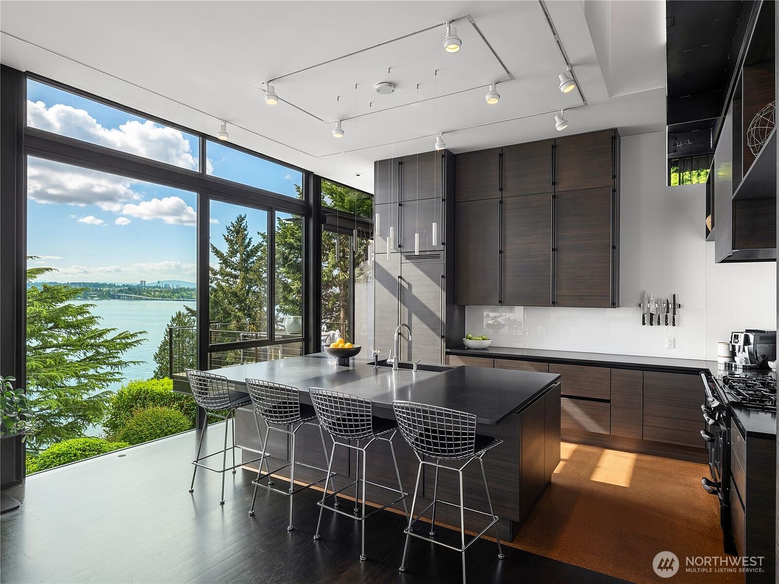This sleek, modern kitchen features dark wood cabinetry and a large central island with a dark countertop and four wire-frame bar stools. The space is defined by expansive floor-to-ceiling windows that provide a panoramic view of the water and surrounding trees, bathing the room in natural light. A track lighting system is mounted on the ceiling, and a magnetic knife strip is mounted on the white backsplash, creating a clean and sophisticated aesthetic.