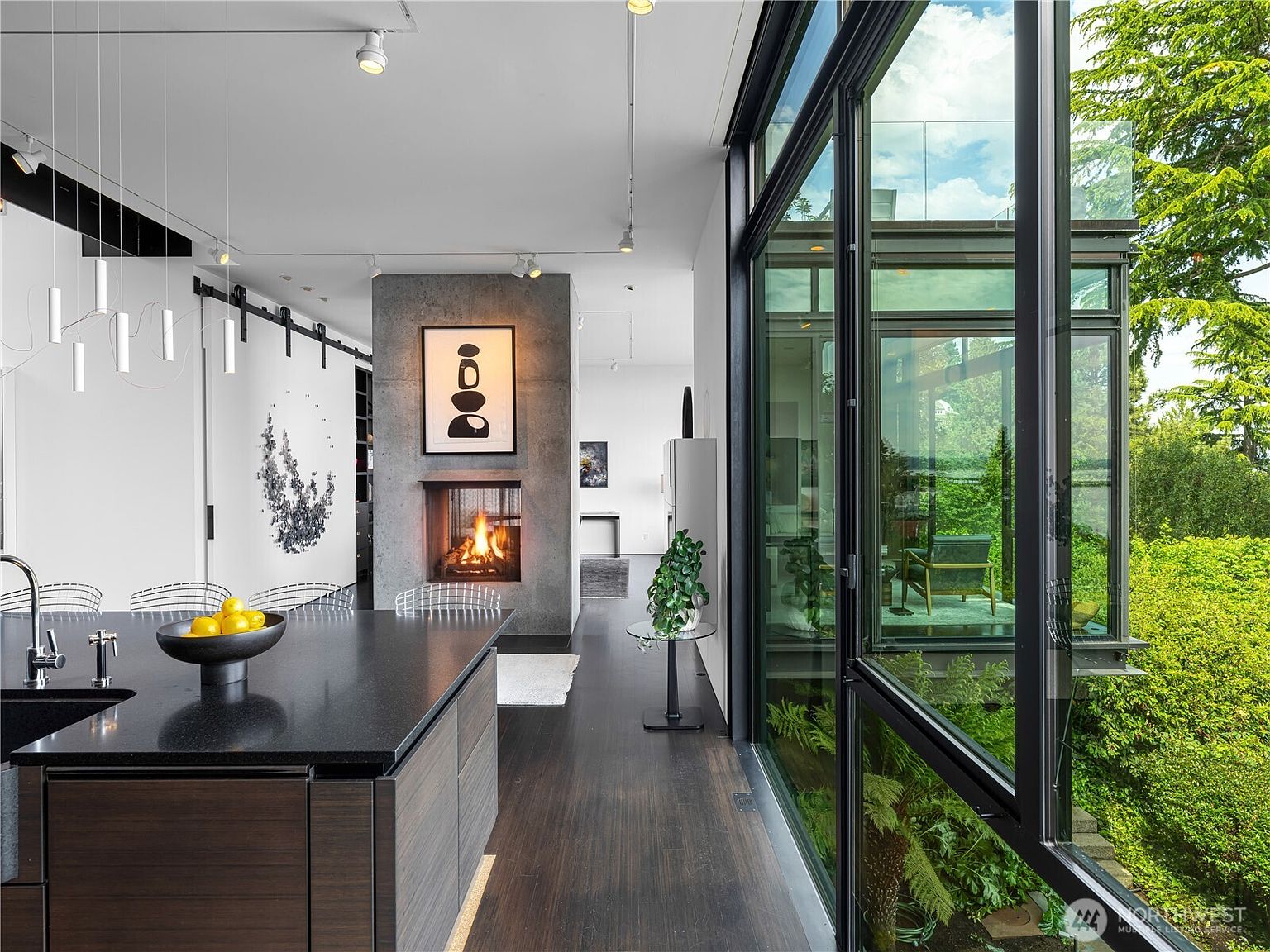 This modern kitchen features a dark island with a bowl of lemons and a sleek faucet, set against a backdrop of dark wood flooring and white walls. A prominent concrete chimney breast houses a fireplace, creating a striking focal point between the kitchen and the adjacent living area. Large glass panels provide an expansive view of the lush greenery outside, blending the interior space with the natural environment.