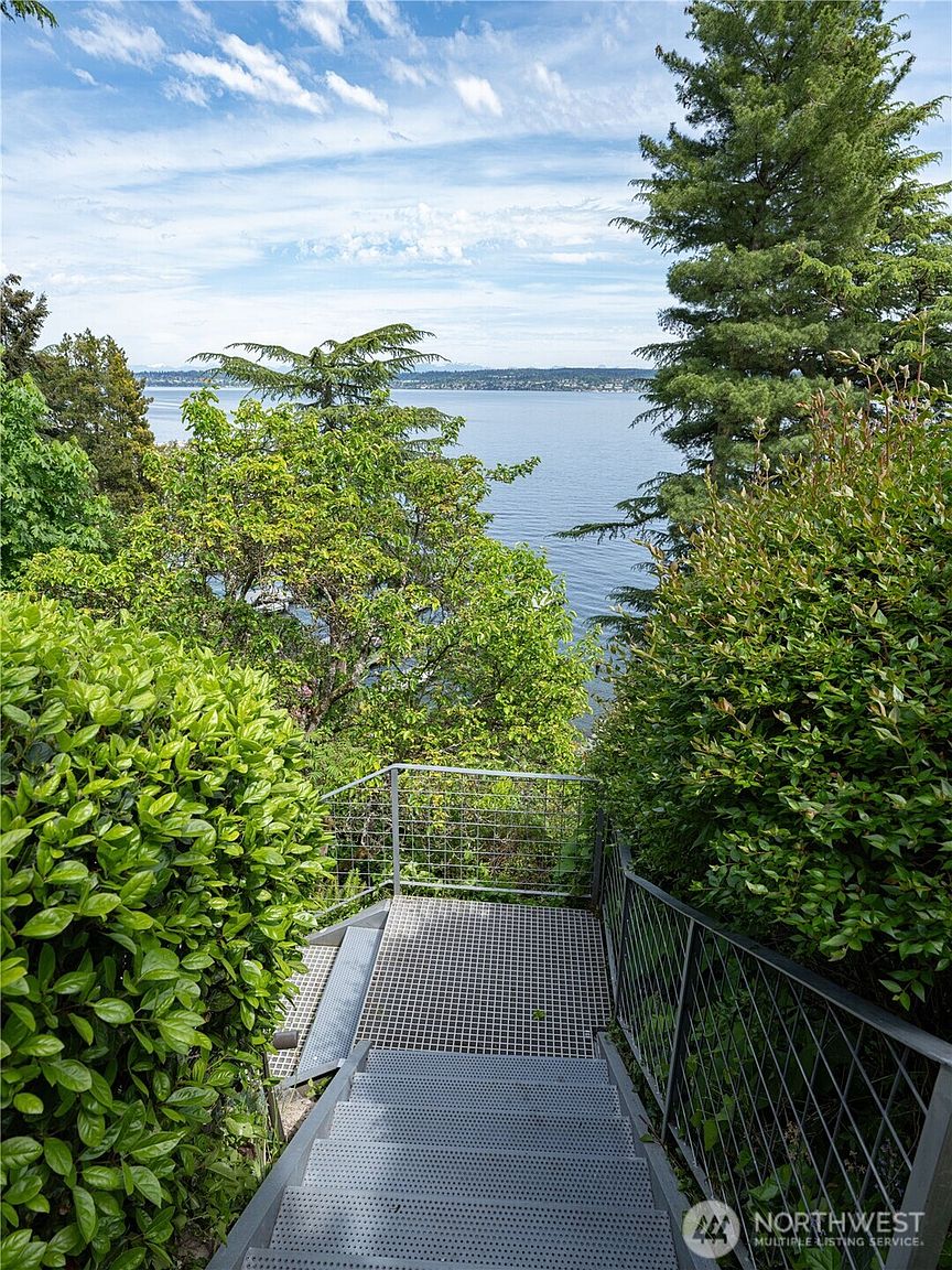 This image captures a set of industrial-style metal stairs leading down toward a scenic waterfront view. The staircase features a durable metal grate design and is enclosed by simple, modern metal railings that provide safety while maintaining an open feel. Lush green trees and foliage frame the view, creating a private and tranquil atmosphere overlooking the expansive body of water.