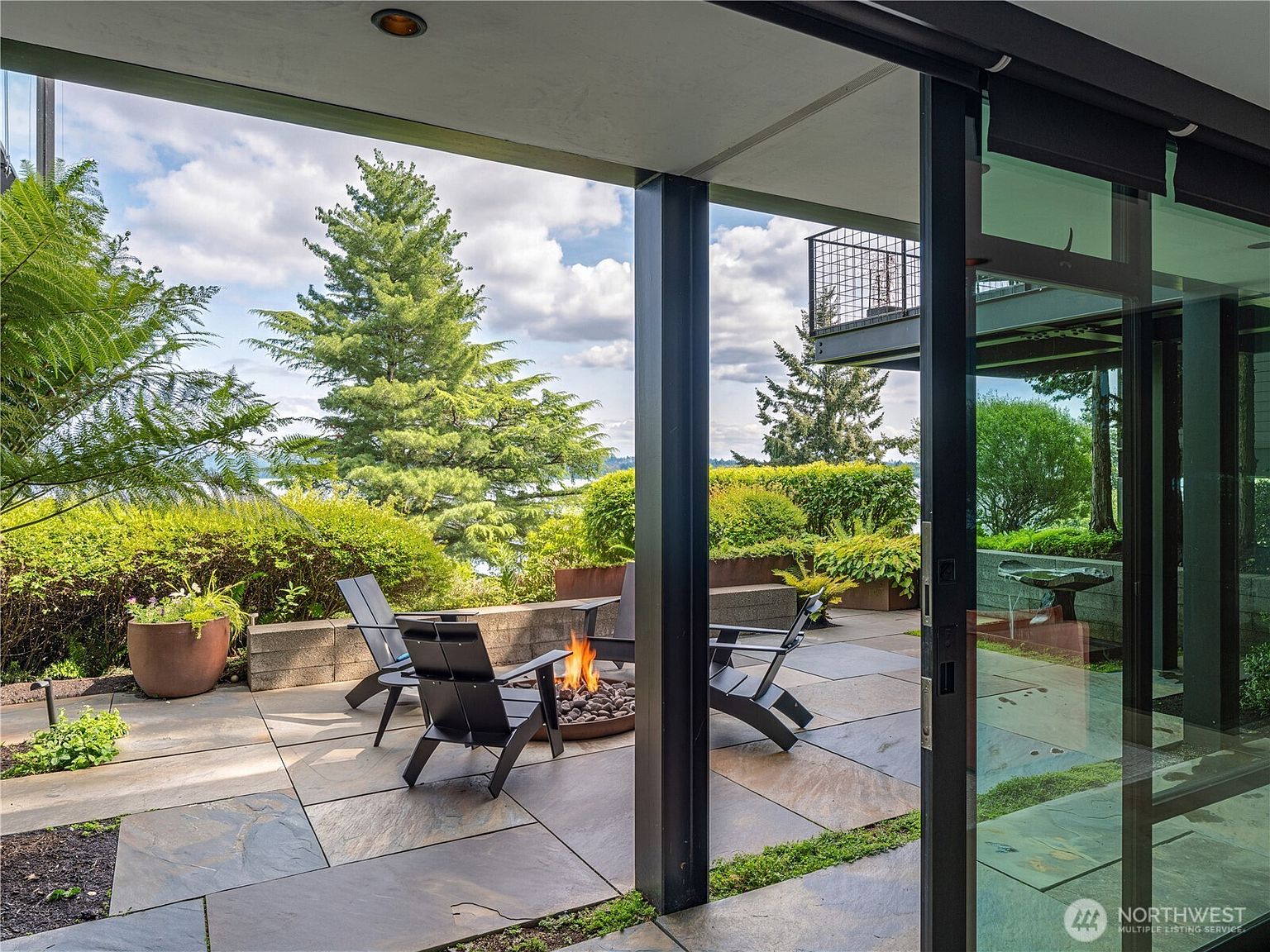 This image captures a modern outdoor patio space viewed from inside through large sliding glass doors. The patio features stone pavers, a circular fire pit surrounded by dark Adirondack-style chairs, and lush landscaping with mature trees in the background. The perspective emphasizes a seamless transition between the indoor living area and the serene, private outdoor retreat.