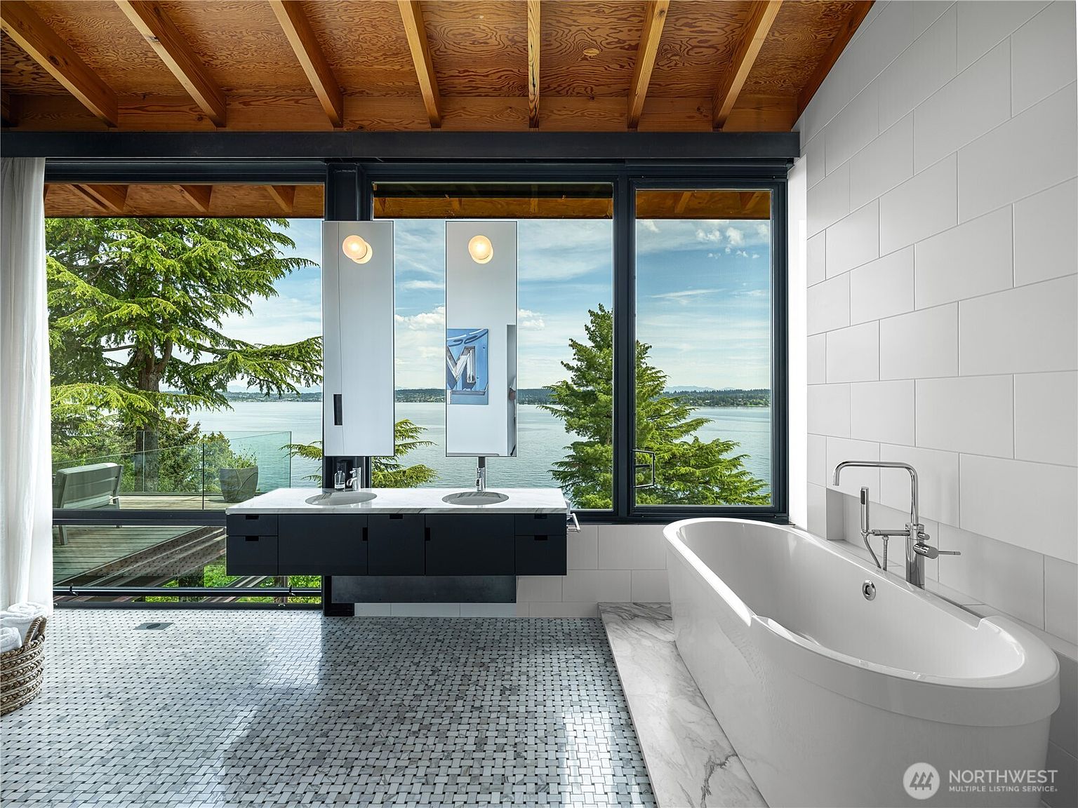 This modern primary bathroom features a floating double vanity with integrated sinks and minimalist mirrors, set against a backdrop of expansive floor-to-ceiling windows that offer a panoramic view of the water and surrounding trees. A sleek, freestanding soaking tub sits on a stone platform, complemented by white tiled walls and a patterned mosaic tile floor. The room is defined by its clean lines, natural wood ceiling beams, and a seamless connection to the outdoor landscape.