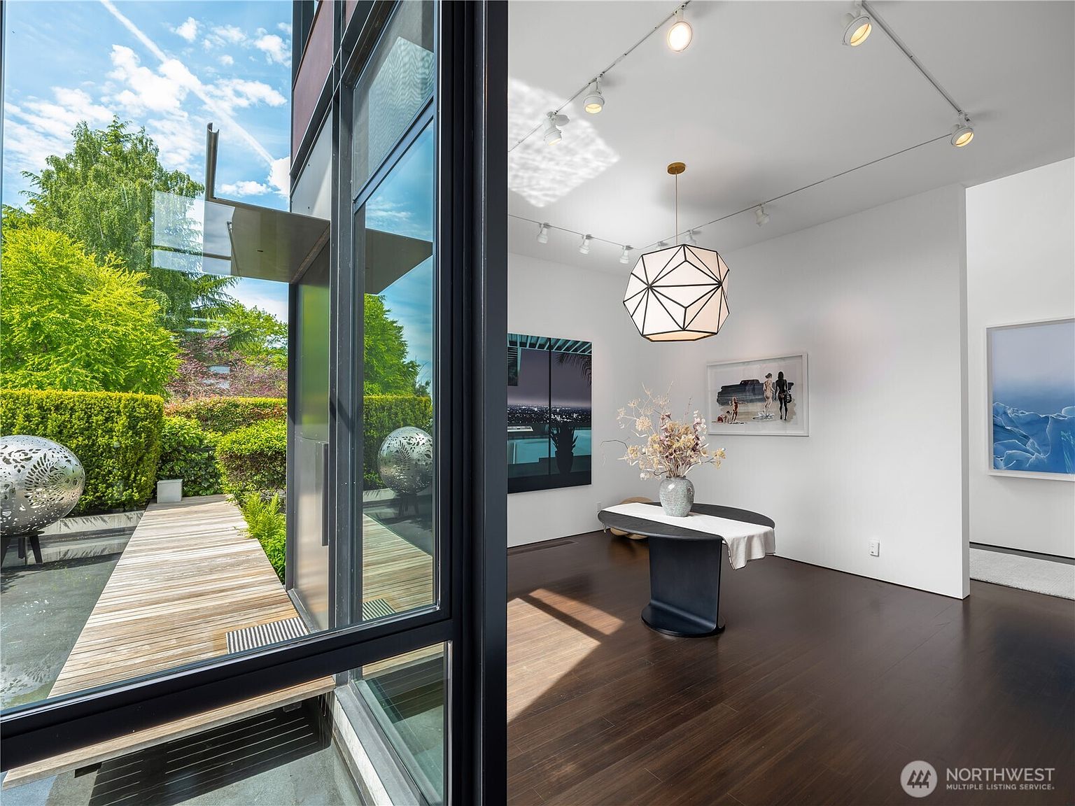 This image captures a seamless transition between an indoor gallery-style foyer and an outdoor wooden deck, separated by large glass panels. The interior features dark wood flooring, a geometric pendant light, and minimalist white walls adorned with contemporary artwork. Outside, a landscaped garden with spherical metal sculptures and lush greenery creates a serene, modern aesthetic.