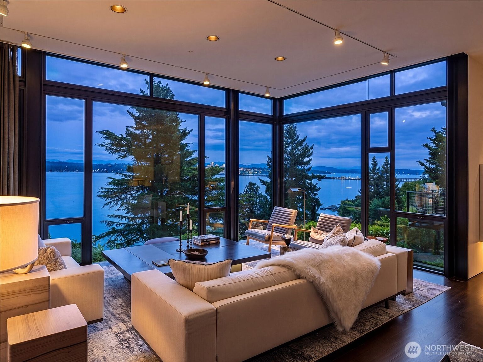 This sophisticated living room features expansive floor-to-ceiling windows that frame a stunning panoramic view of the water and distant shoreline at dusk. The space is furnished with a plush, light-toned sectional sofa adorned with a faux-fur throw, two modern accent chairs, and a large, dark-toned coffee table. Recessed track lighting illuminates the room, creating a warm and inviting atmosphere against the cool blue tones of the evening sky outside.