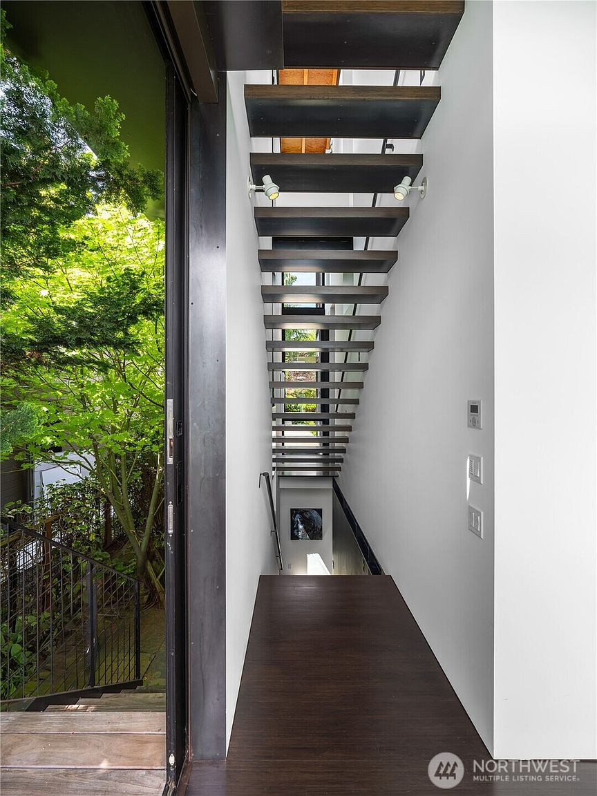 This interior view captures a striking, modern staircase with floating dark wood treads that ascend along a crisp white wall. To the left, a large glass door opens to a lush, green outdoor area, creating a seamless connection between the indoor space and nature. The perspective looks down the stairwell, highlighting the minimalist design and the clean lines of the architectural structure.