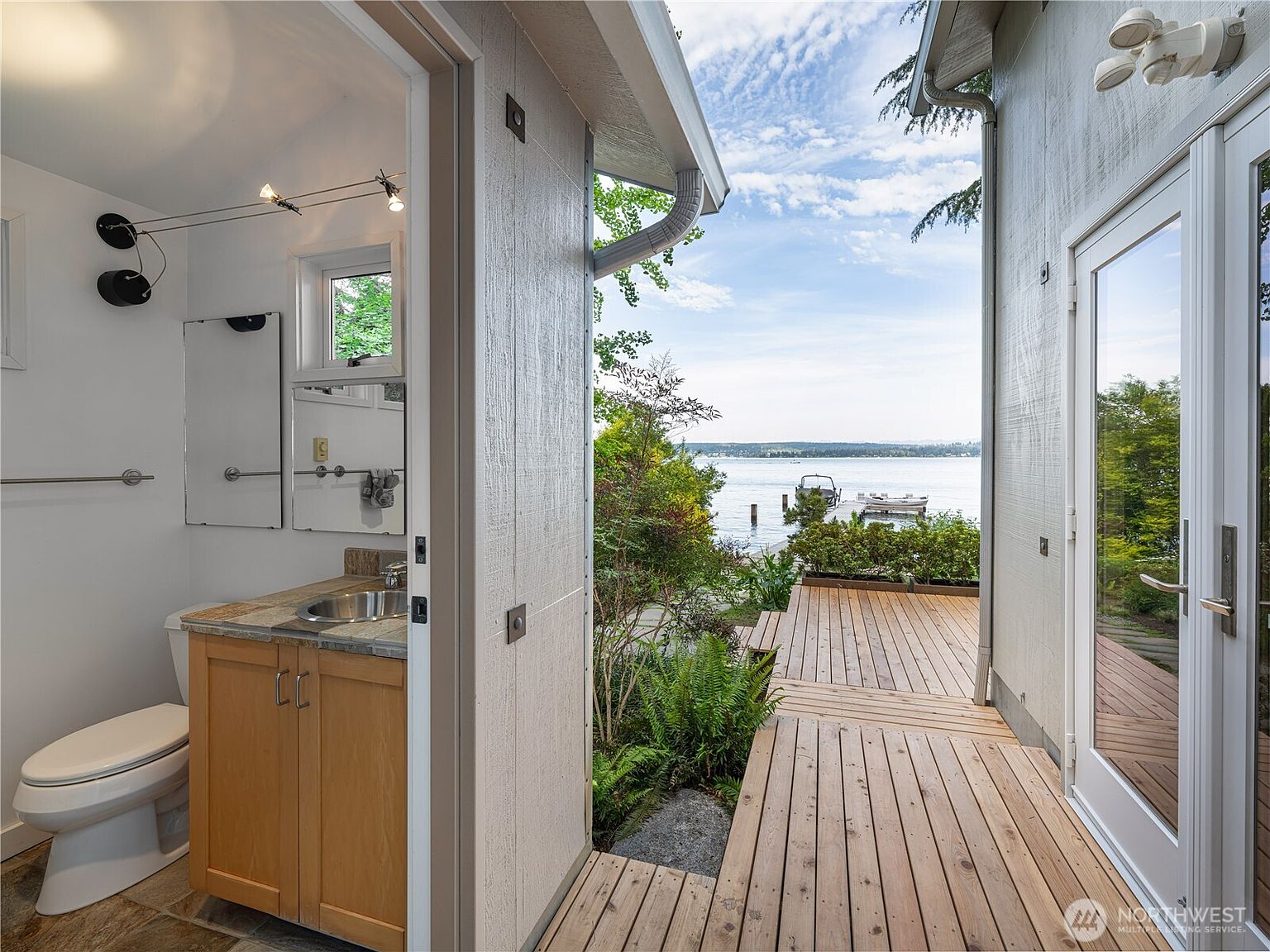 This image captures a unique transition space, showing a compact bathroom interior opening directly onto a wooden deck overlooking a serene waterfront. The bathroom features light wood cabinetry, a metallic sink, and a toilet, while the exterior deck leads the eye toward a calm lake with a private dock and boats in the distance. The perspective is framed from inside the doorway, highlighting the seamless connection between indoor utility and outdoor leisure.