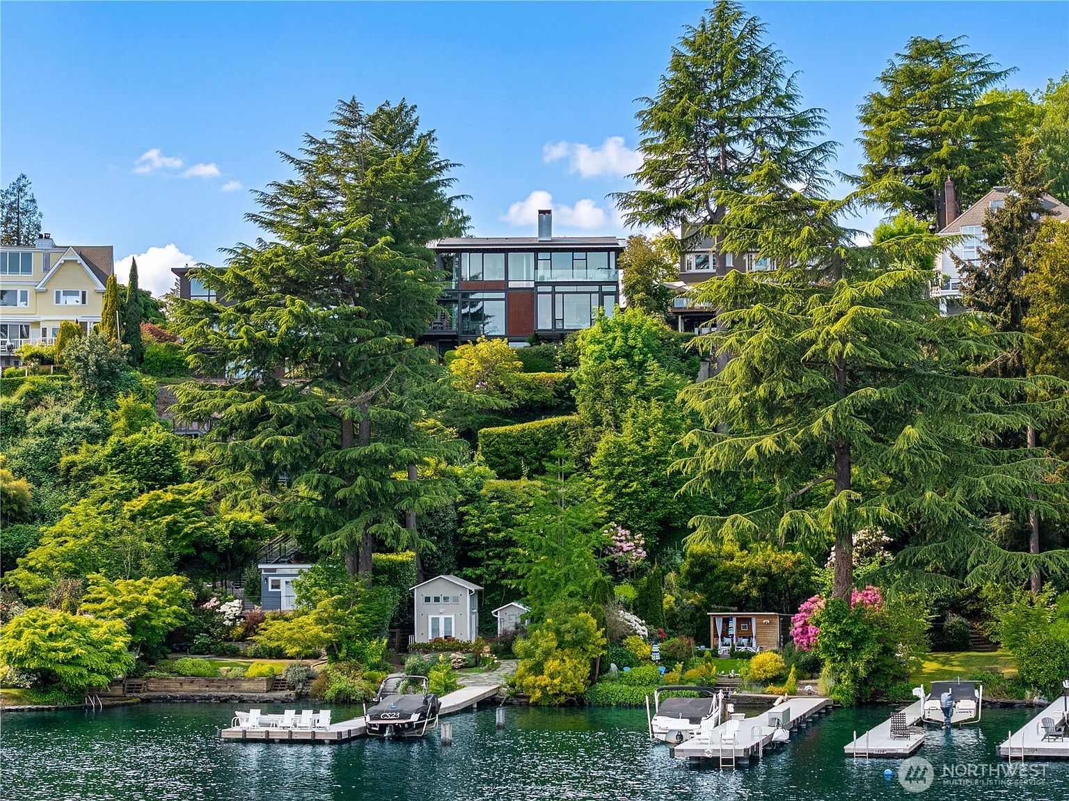 This waterfront property features a modern, multi-level home nestled on a lush, tree-covered hillside overlooking a serene lake. The residence showcases contemporary architecture with large windows and expansive decks, while the shoreline includes private docks and small outbuildings. The perspective is captured from the water, emphasizing the home's elevated position and integration into the natural landscape.
