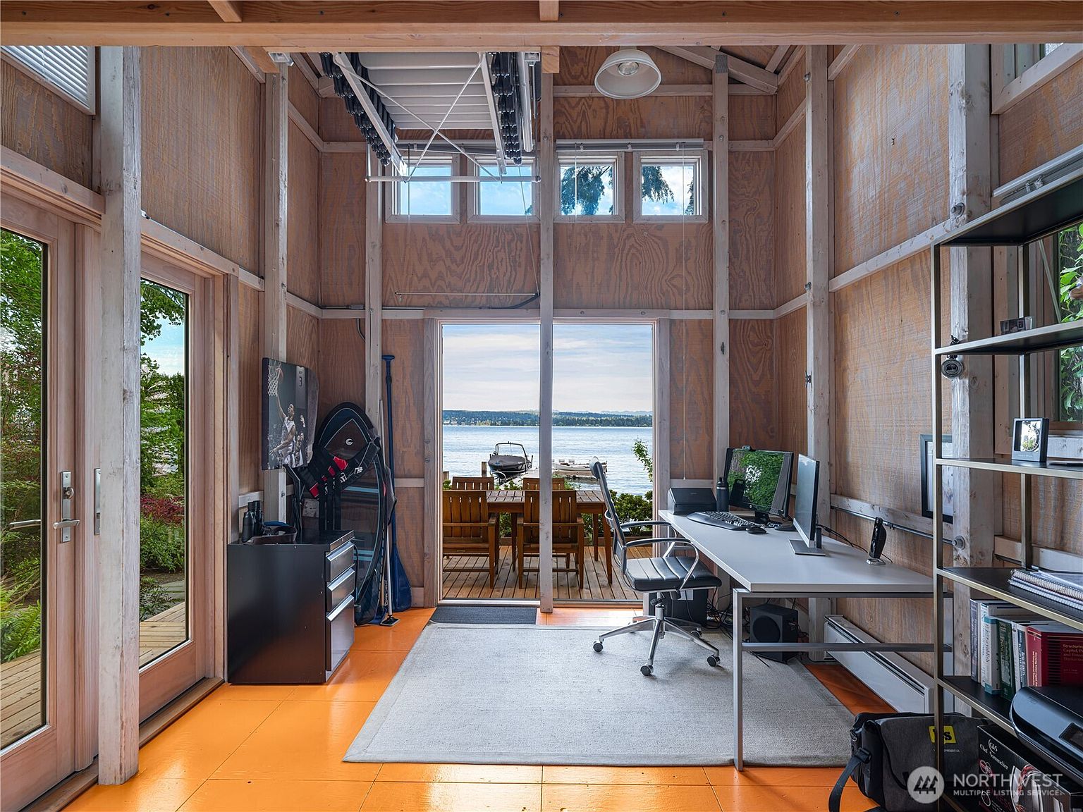 This bright home office features a unique, rustic aesthetic with exposed wood-paneled walls and a vibrant orange floor. A large desk setup is positioned to face a set of glass doors that open onto a deck overlooking a serene lake, providing a picturesque view. The room is equipped with a modern office chair, a shelving unit, and storage cabinets, creating a functional workspace that blends indoor comfort with natural scenery.