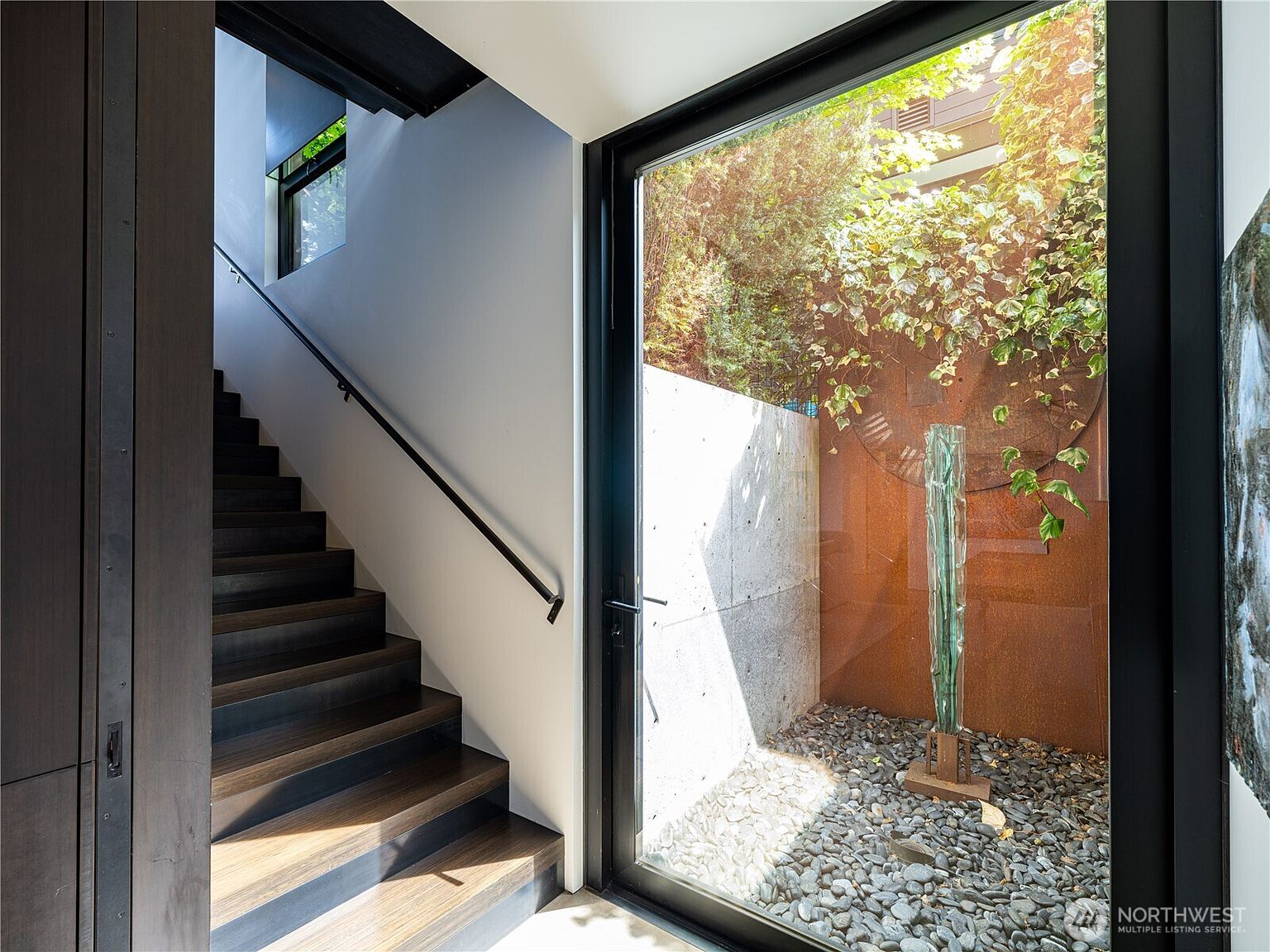 This interior view captures a modern staircase with dark wood treads leading upward, contrasted by a minimalist white wall and a sleek black handrail. To the right, a large glass panel provides a view into a private, enclosed courtyard featuring a sculptural glass art piece, a rusted metal wall, and a bed of river stones. The composition highlights a sophisticated blend of architectural materials and a seamless connection between the indoor staircase and the outdoor light-filled atrium.