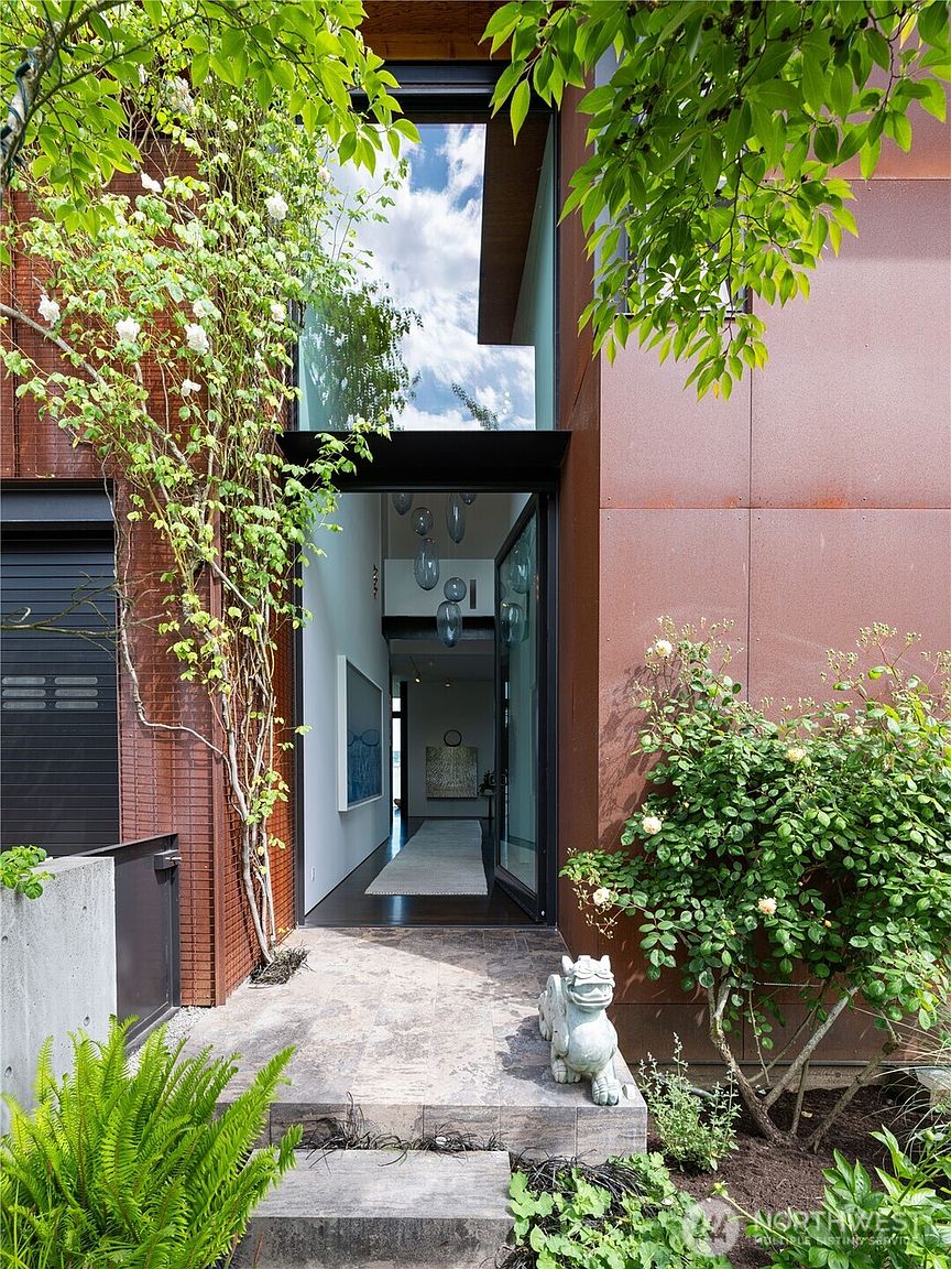 This striking modern entryway features a tall, glass-paneled door framed by rusted metal siding and a climbing rose vine. A stone-tiled path leads to the entrance, flanked by lush greenery and a decorative stone statue. The perspective looks directly into a bright, minimalist hallway adorned with hanging pendant lights and contemporary artwork.