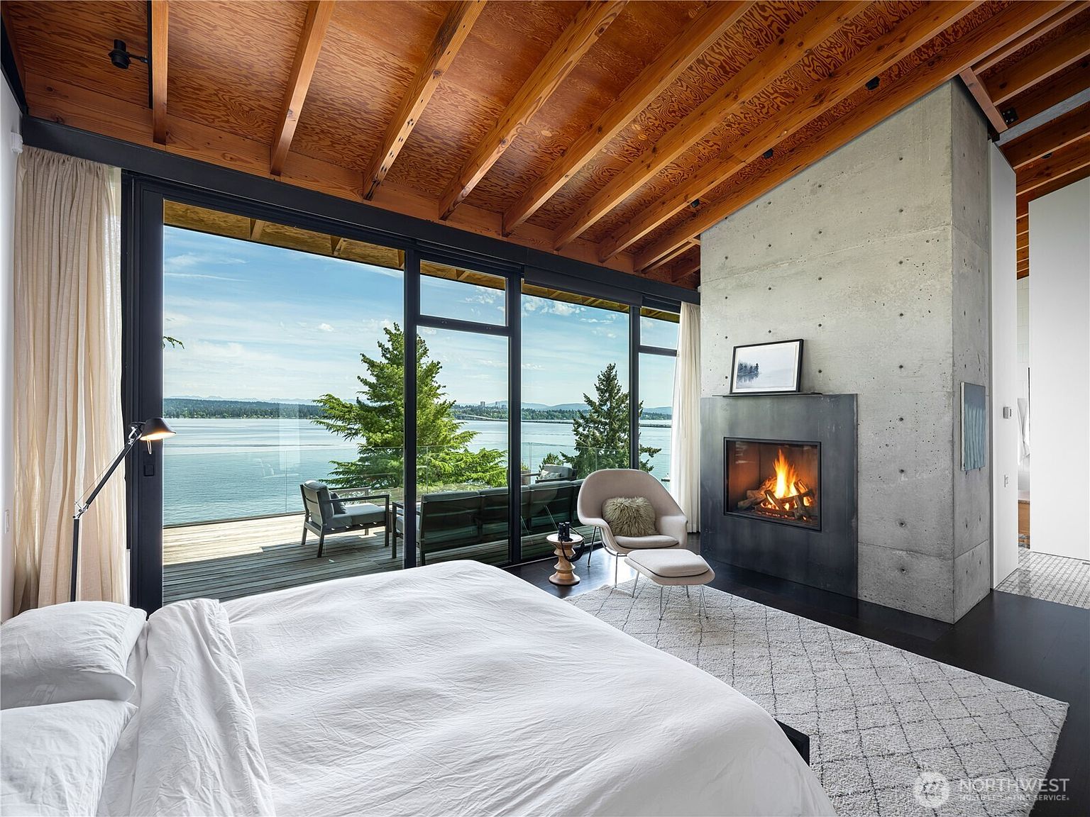 This primary bedroom features a striking concrete chimney breast housing a fireplace, set against a backdrop of expansive floor-to-ceiling glass walls that offer panoramic water views. The room is defined by its warm wood-beamed ceiling, dark flooring, and a minimalist aesthetic, complemented by a comfortable lounge chair and ottoman. A large white bed occupies the foreground, while the seamless transition to the outdoor deck enhances the sense of indoor-outdoor living.