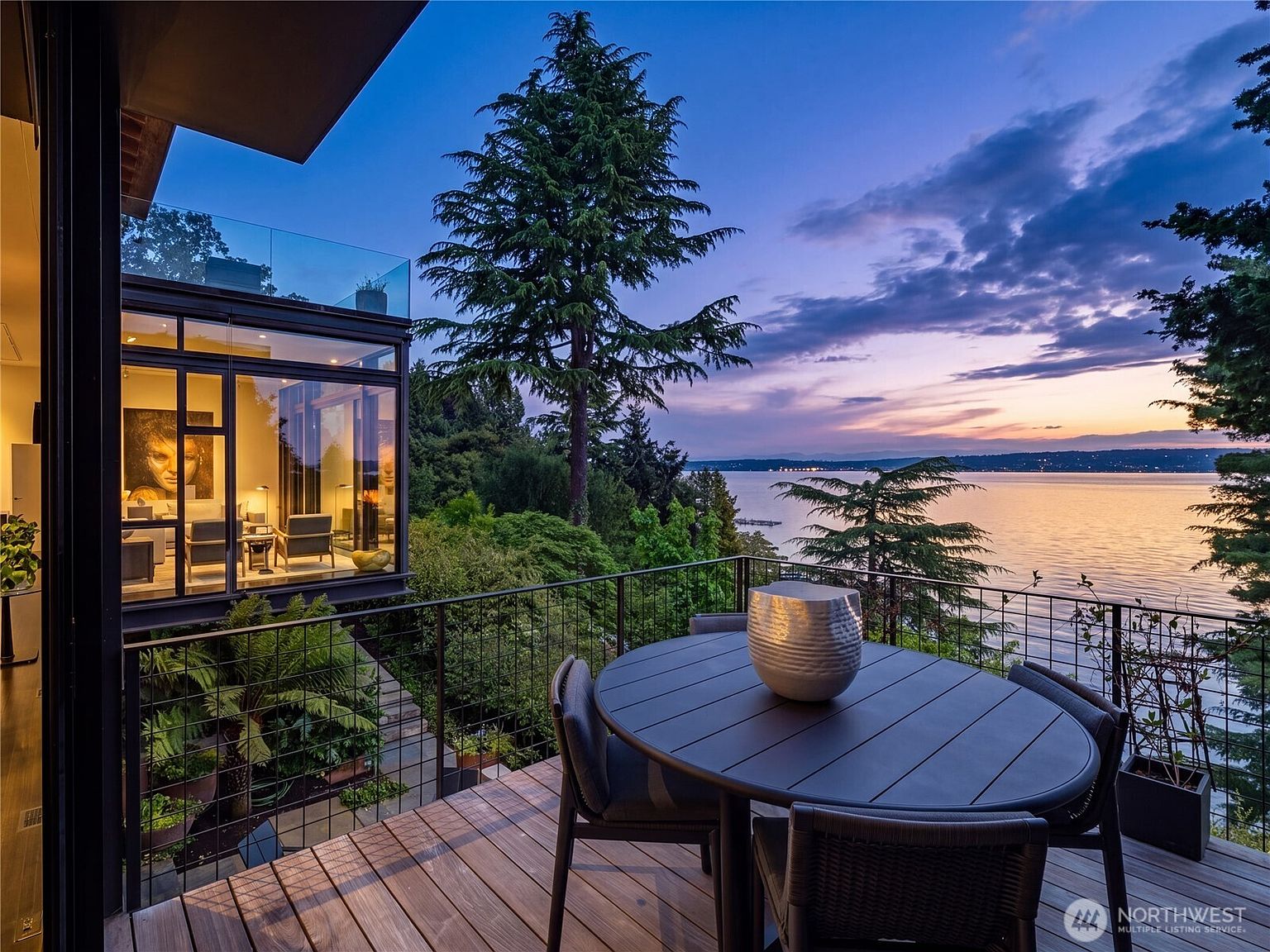 This elevated deck offers a stunning, cinematic perspective of a tranquil waterfront at dusk, framed by lush trees and a modern architectural structure. A circular dining table with dark chairs sits in the foreground, while the adjacent interior space is visible through expansive glass walls, showcasing a contemporary living area. The scene captures a sophisticated blend of indoor-outdoor living, highlighted by the soft, ambient glow of the interior lighting against the twilight sky.