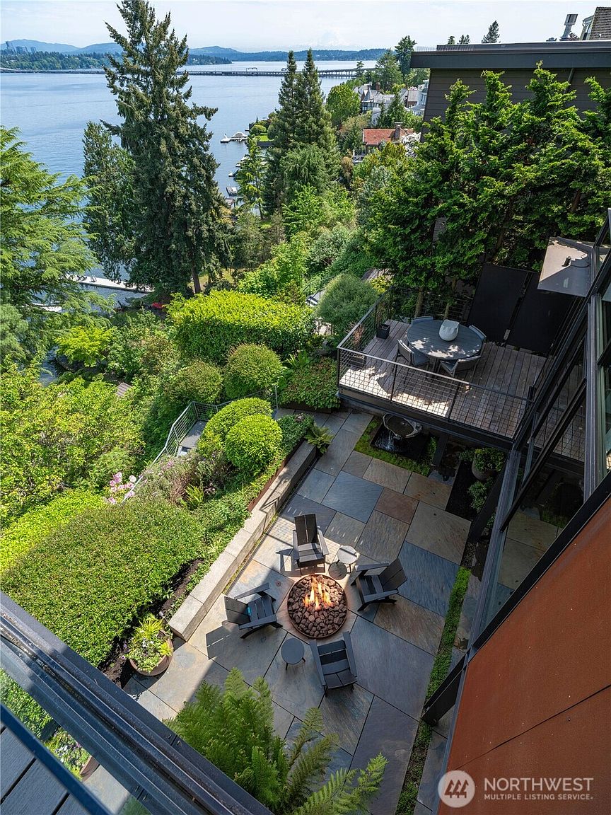 This high-angle shot captures a multi-level outdoor living space featuring a stone-paved patio with a central fire pit surrounded by dark chairs. Above the patio, a wooden deck provides an elevated dining area, while lush greenery and mature trees frame a stunning view of the lake and distant bridge. The perspective emphasizes the seamless integration of the home's architecture with the natural, sloped landscape.