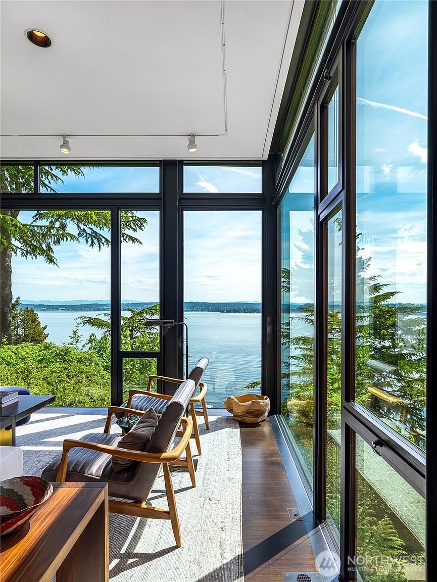 This modern living space features expansive floor-to-ceiling windows that offer panoramic views of the water and surrounding trees. The room is furnished with two mid-century modern style chairs and a low-profile table, set against a neutral area rug on dark wood flooring. The minimalist design emphasizes the connection between the interior and the natural landscape outside.