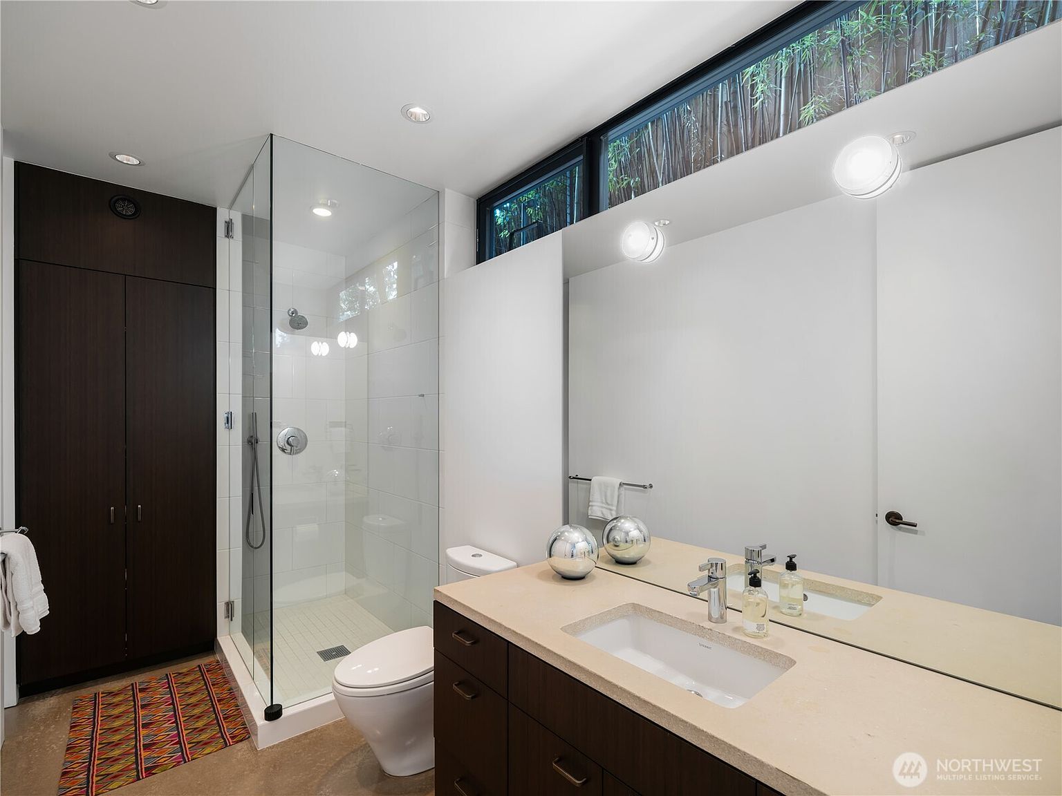 This modern bathroom features a sleek vanity with a light-toned countertop and dark cabinetry, paired with a large wall-to-wall mirror. A glass-enclosed walk-in shower with white tile walls sits adjacent to a white toilet, while a clerestory window above the mirror allows natural light to filter in from the outdoors. The space is finished with a patterned rug on the floor, creating a clean and contemporary aesthetic.