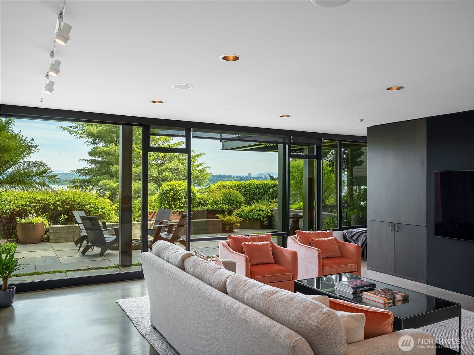 This modern living room features a neutral-toned sofa and two plush, salmon-colored armchairs arranged around a glass-topped coffee table. The space is defined by expansive floor-to-ceiling windows that provide a seamless transition to an outdoor patio and lush greenery beyond. A dark, sleek media wall contrasts with the light interior, while recessed lighting and track lights illuminate the contemporary living area.