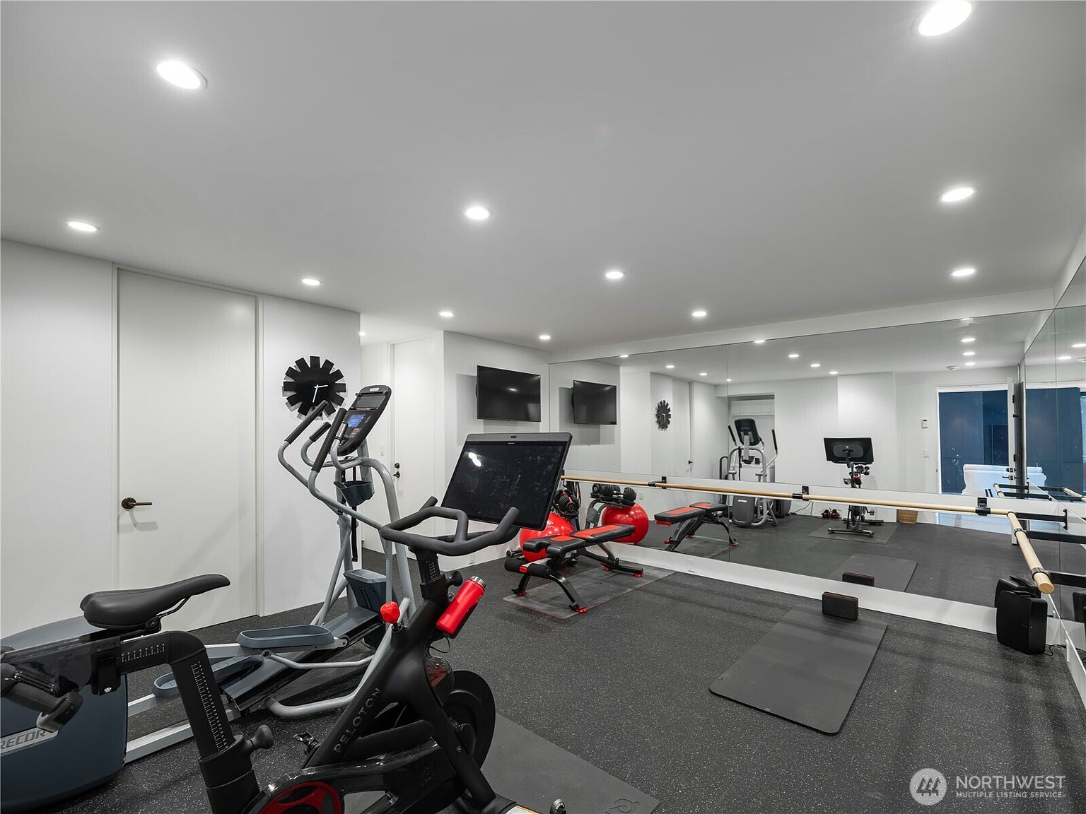 This dedicated home gym features a clean, minimalist aesthetic with white walls and recessed lighting. The space is equipped with a Peloton bike, an elliptical machine, a weight bench, and a ballet barre mounted against a large wall mirror that enhances the room's depth. A durable, dark speckled rubber floor provides a functional surface for various workout routines.