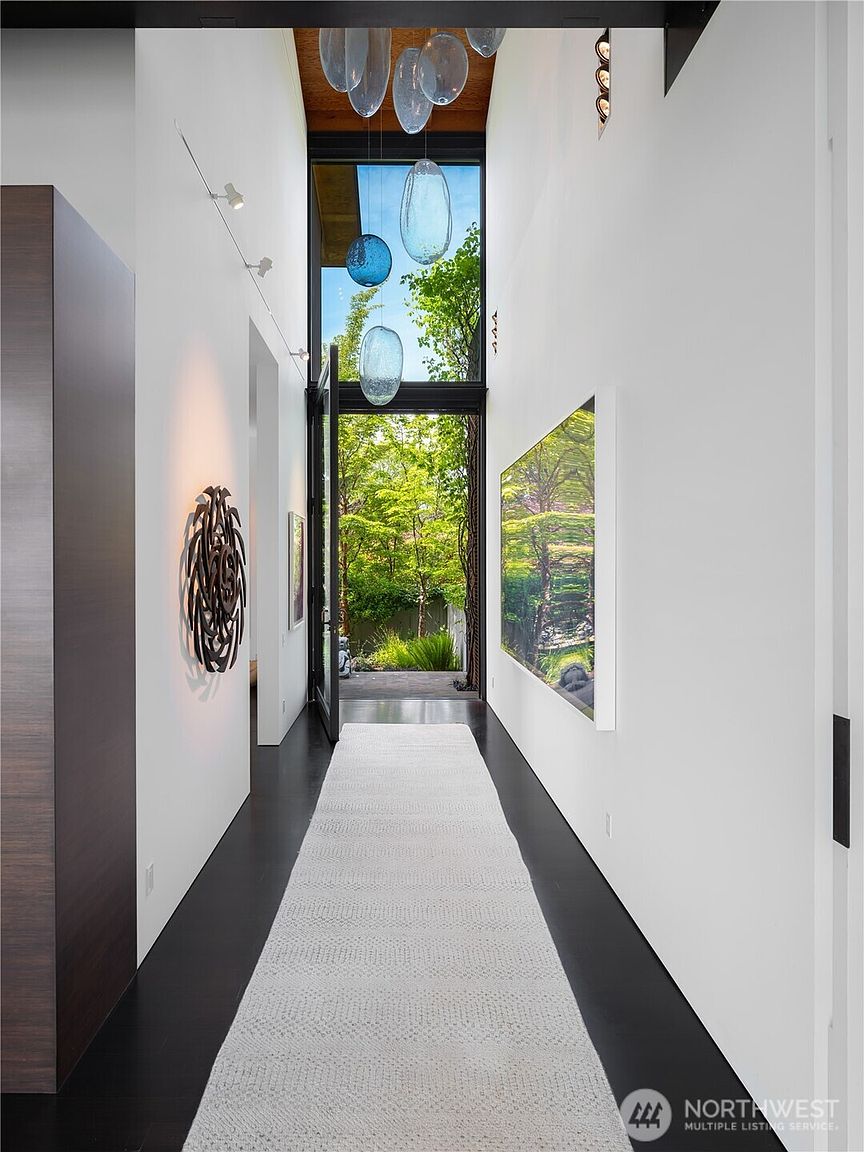 This striking, high-ceilinged hallway features dark flooring contrasted by crisp white walls and a long, textured runner rug. A dramatic glass-walled entryway at the end of the hall provides a seamless transition to the lush greenery outside, while a collection of suspended, organic-shaped glass pendants adds an artistic focal point. The space is further enhanced by a large, abstract wall sculpture on the left and a expansive landscape art piece on the right, creating a sophisticated and gallery-like atmosphere.