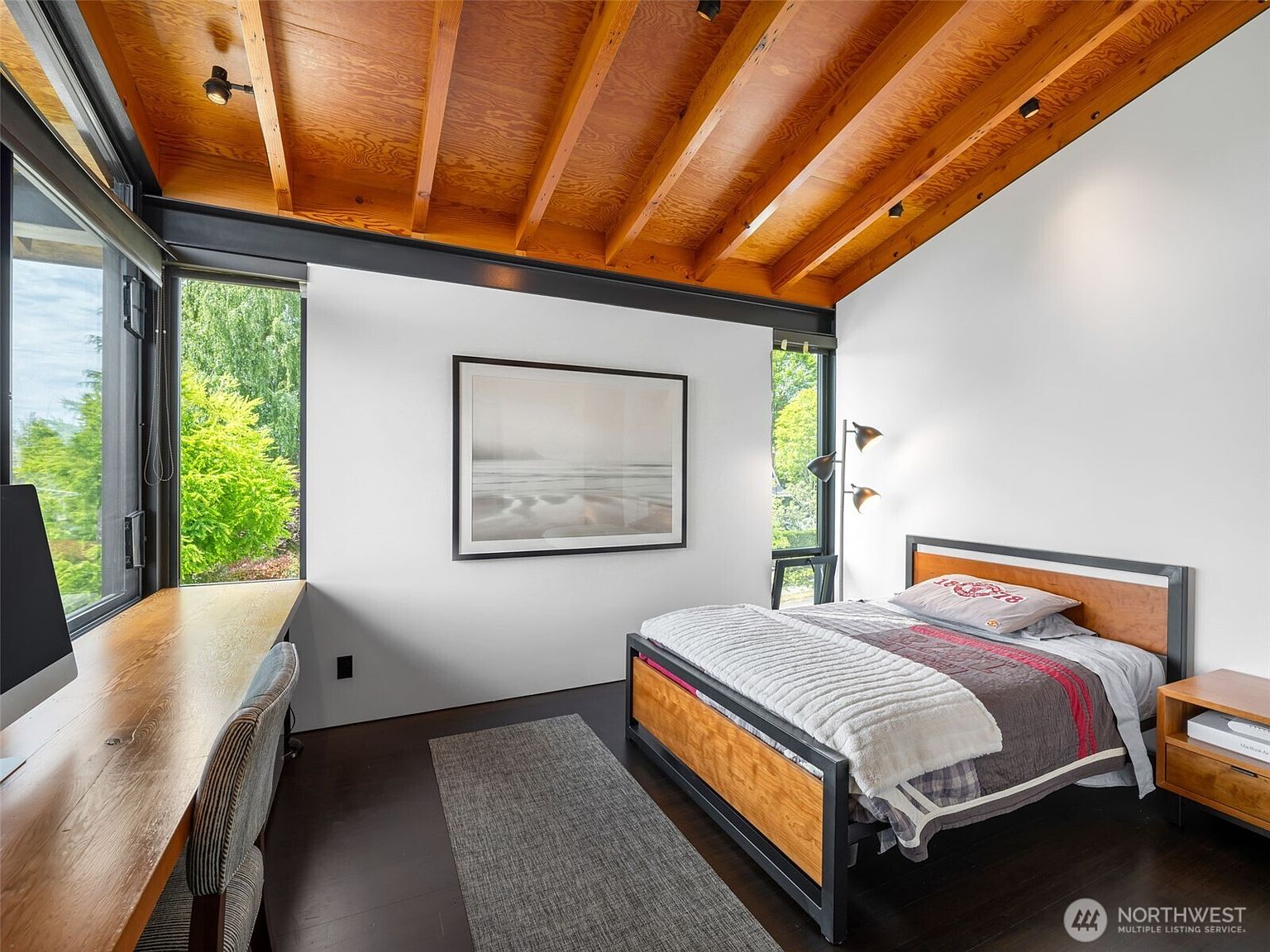 This modern bedroom features a striking vaulted ceiling with exposed wood beams and a dark wood floor. A built-in desk runs along the large windows, providing a bright workspace, while a metal-framed bed with wood accents serves as the focal point. The room is decorated with minimalist art and a floor lamp, creating a clean, contemporary aesthetic with a view of the surrounding greenery.
