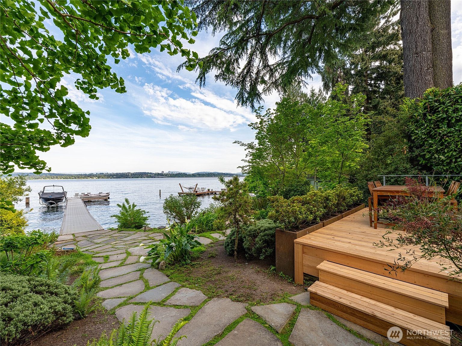 This scenic outdoor space features a natural stone pathway leading toward a private dock on a calm lake. A tiered wooden deck with an outdoor dining set provides a comfortable vantage point for enjoying the waterfront views, surrounded by lush greenery and mature trees. The perspective captures the serene transition from the landscaped garden to the expansive open water.