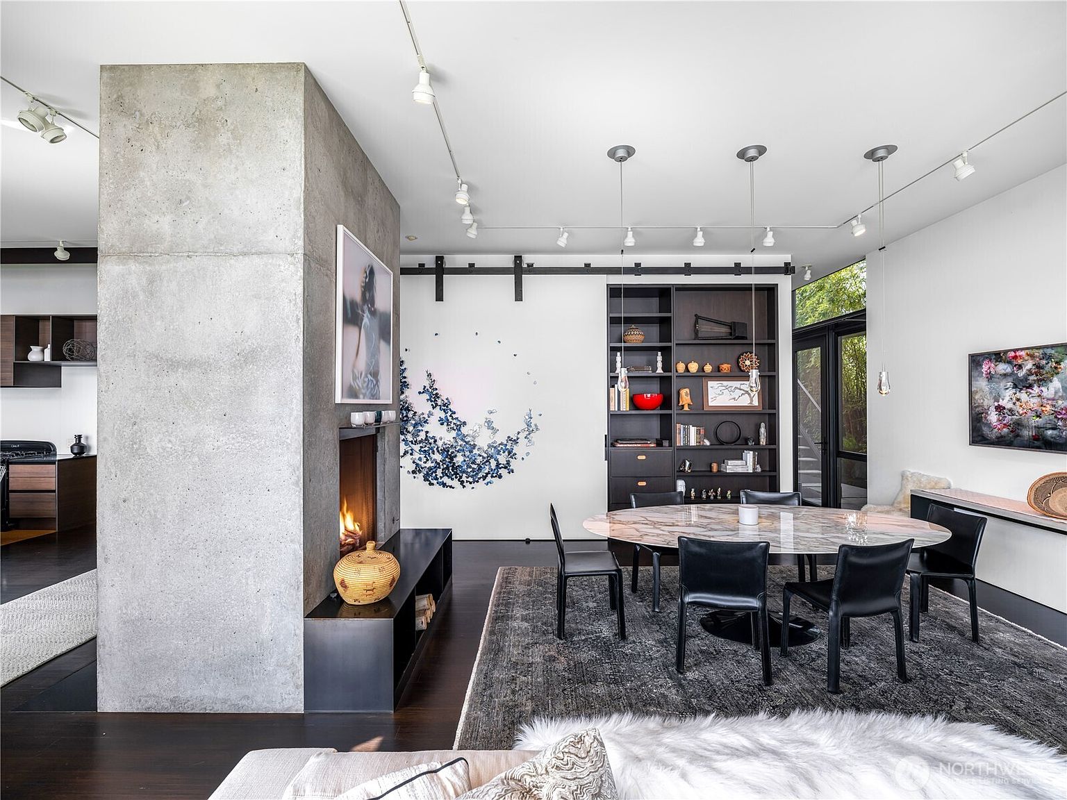 This sophisticated dining area features a large oval table with a stone-patterned top surrounded by black chairs, set upon a textured area rug. A prominent concrete chimney breast anchors the space, housing a fireplace and a piece of art, while a dark built-in shelving unit provides elegant storage. The room is illuminated by track lighting and modern pendant fixtures, creating a refined, contemporary atmosphere with a view into an adjacent kitchen.