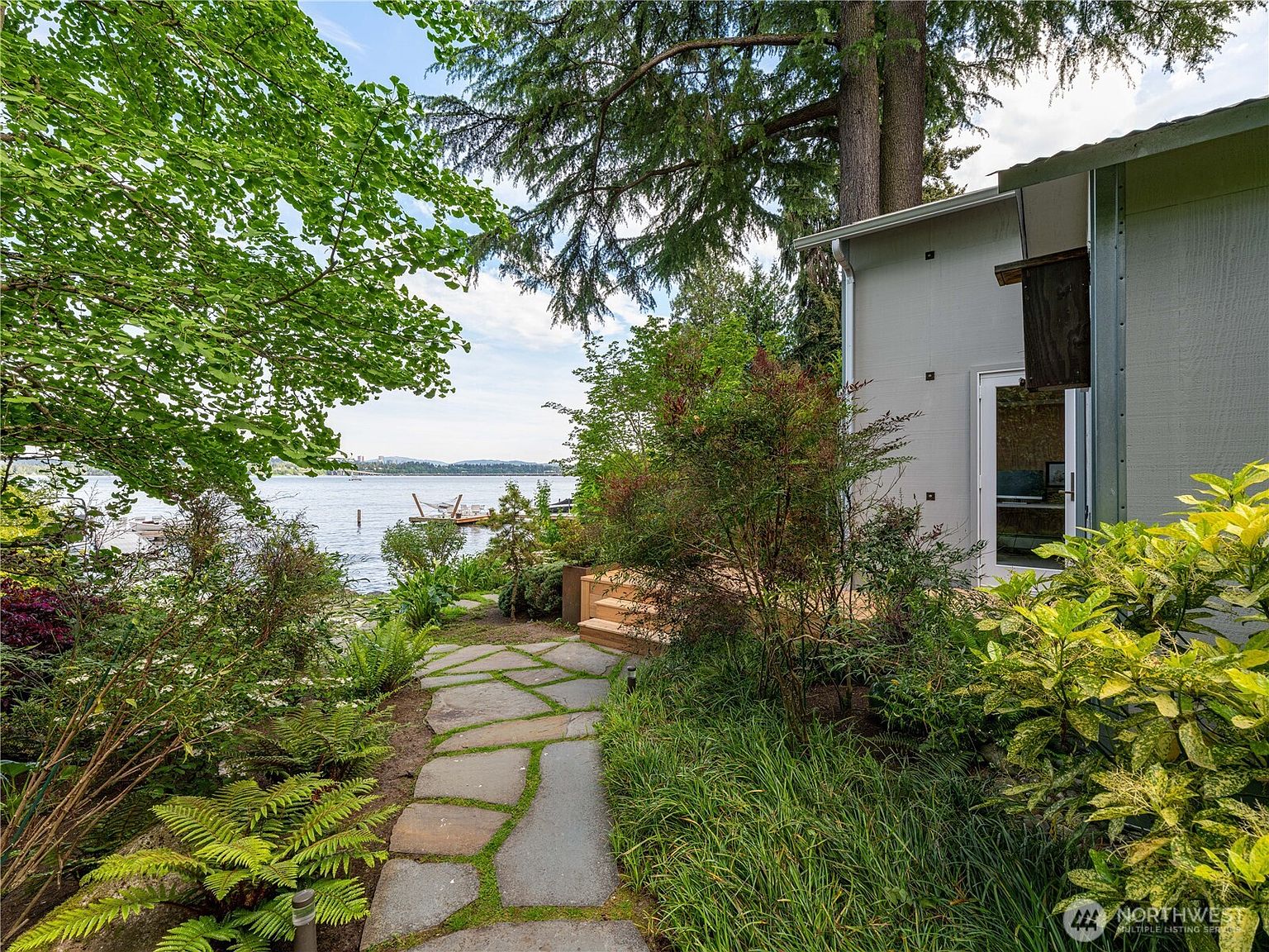 This serene outdoor space features a natural stone pathway winding through lush, mature landscaping toward a wooden deck and a waterfront view. The scene includes a side view of a light-colored home with a glass door, framed by vibrant green foliage and tall trees. The overall atmosphere is tranquil and private, highlighting the seamless connection between the home's architecture and the natural lakeside environment.