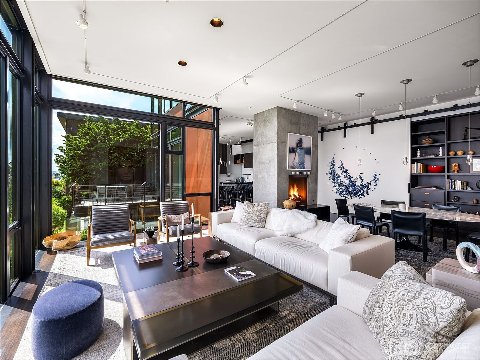 This sophisticated living room features a contemporary open-concept design with expansive floor-to-ceiling windows that provide a seamless connection to the outdoor deck. The space is anchored by a large white sectional sofa, a dark metal coffee table, and a central concrete chimney breast housing a warm fireplace. A dining area with a dark bookshelf and modern seating is visible in the background, creating a cohesive and luxurious atmosphere.