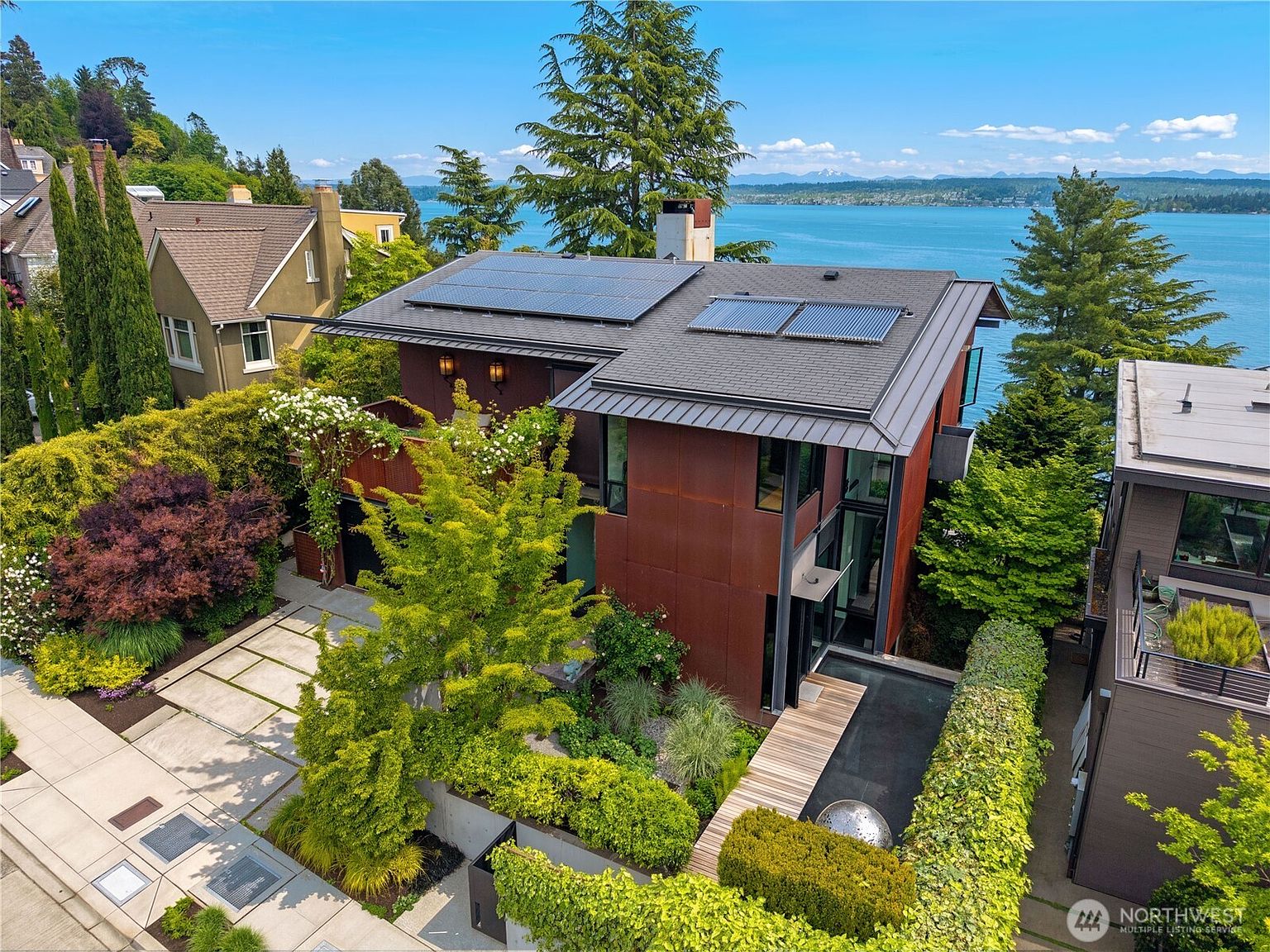 This high-angle aerial view captures a contemporary home with a distinctive rust-colored metal facade and a dark, multi-level roof equipped with solar panels. The property features a landscaped front garden with a wooden walkway leading to the entrance, set against a stunning backdrop of a large lake and distant mountains. The architectural style is modern and industrial, emphasizing clean lines and a seamless integration with the surrounding lush greenery.