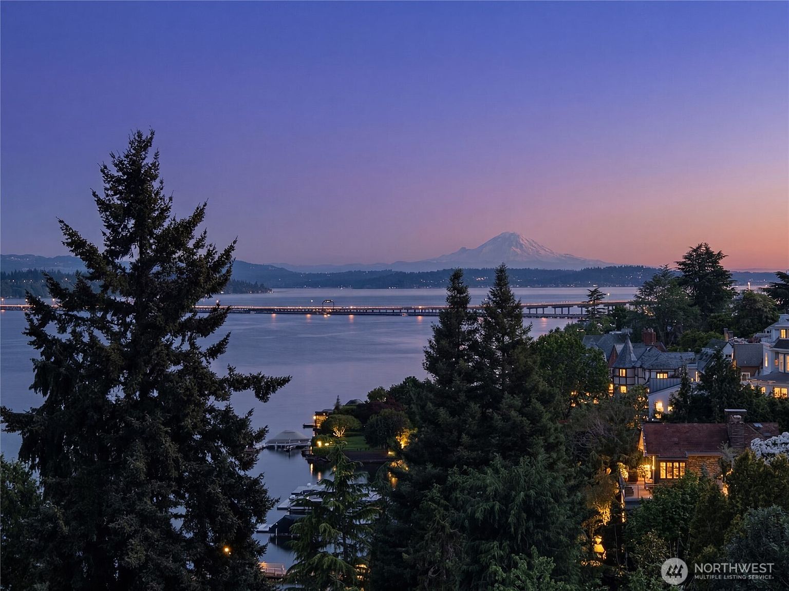 This elevated, wide-angle view captures a stunning twilight scene overlooking a calm body of water with a prominent bridge stretching across the horizon. In the distance, a majestic, snow-capped mountain stands against a soft purple and blue gradient sky. The foreground features lush, dark evergreen trees framing the view, with glimpses of residential homes nestled along the shoreline, illuminated by warm, inviting lights.