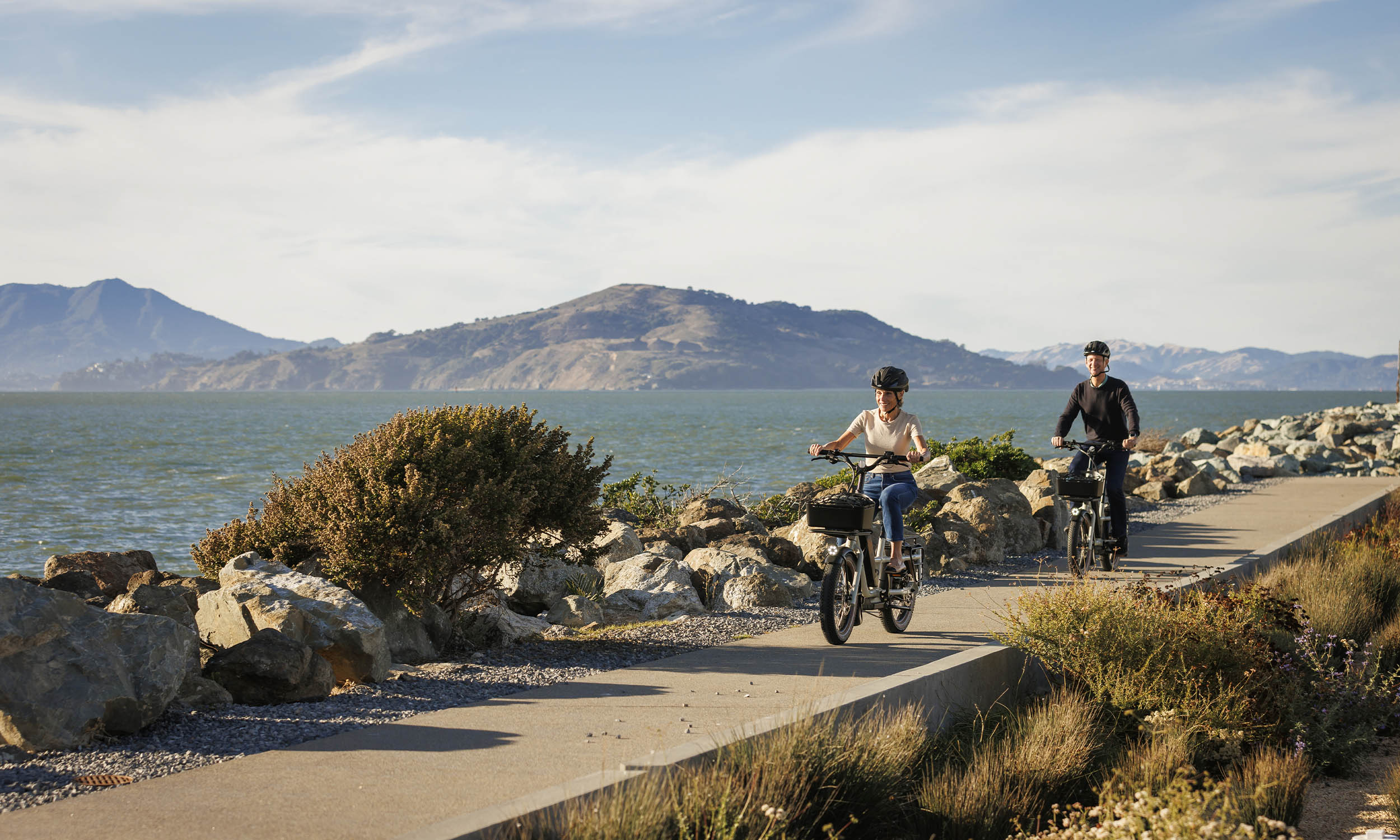 This image showcases a scenic side view featuring two individuals riding electric bikes along a paved path adjacent to a body of water. The backdrop includes rolling hills and a clear sky, creating a serene and inviting atmosphere. The path is lined with rocks and vegetation, adding to the natural beauty of the location.