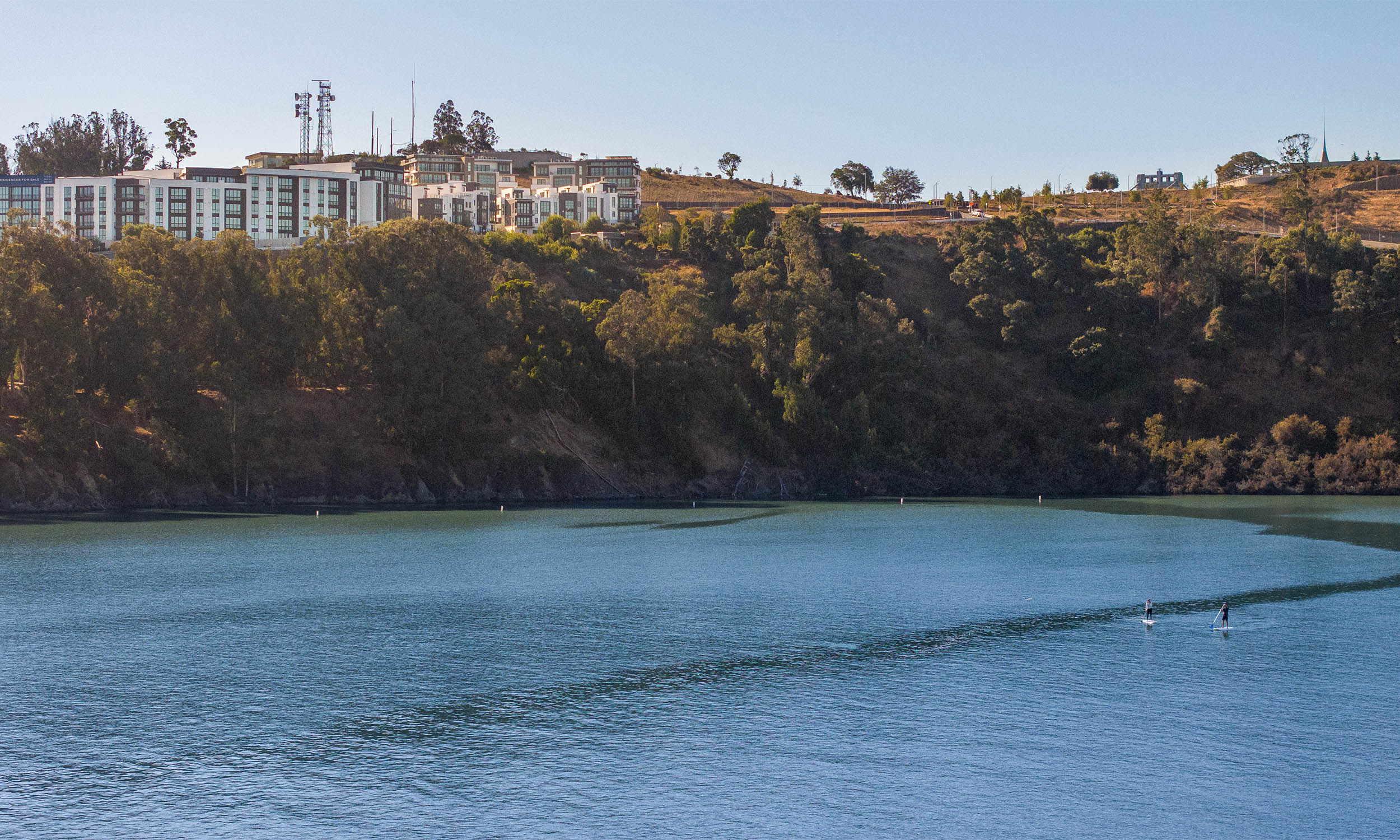 This aerial view showcases a modern residential complex situated on a hillside overlooking a serene body of water. The architecture features clean lines and a contemporary design, with multiple buildings arranged to maximize views. Two paddleboarders are visible on the water, adding a sense of recreation and tranquility to the scene.