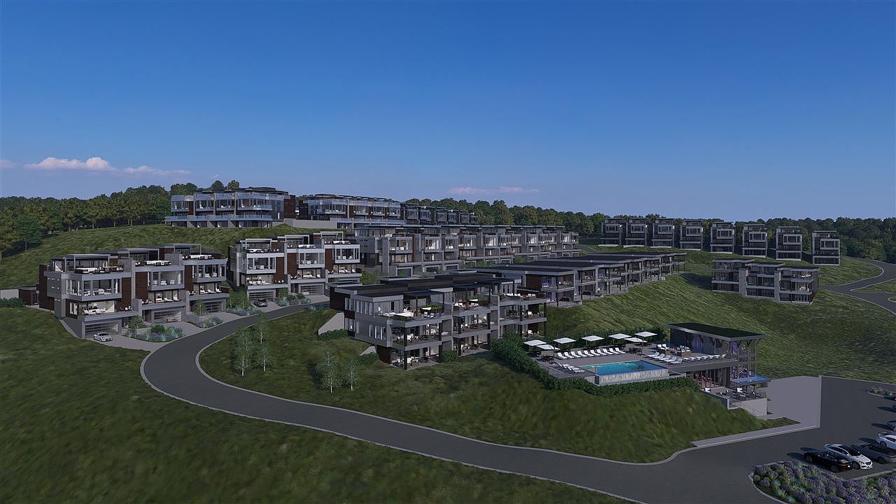 This aerial view showcases a modern residential complex nestled on a green hillside under a clear blue sky. The architecture features clean lines, flat roofs, and a mix of gray and brown tones, with multiple units boasting balconies and large windows. A winding road connects the buildings, and a central amenity area includes a pool and parking lot, creating an impression of upscale community living.