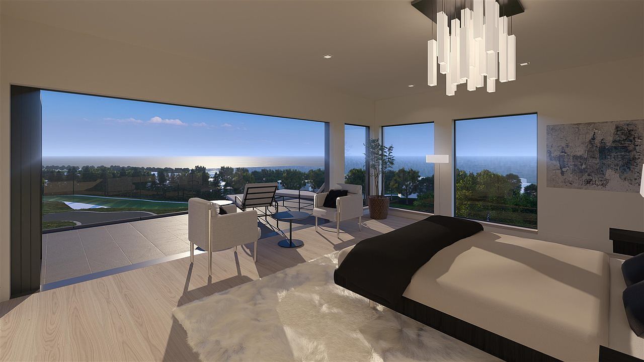 This is a primary bedroom featuring a modern design with a large window offering a scenic view of the ocean and landscape. The room includes a bed with a black and beige color scheme, a white fluffy rug, and a seating area with chairs and a small table. A contemporary chandelier hangs from the ceiling, adding to the room's elegant ambiance.