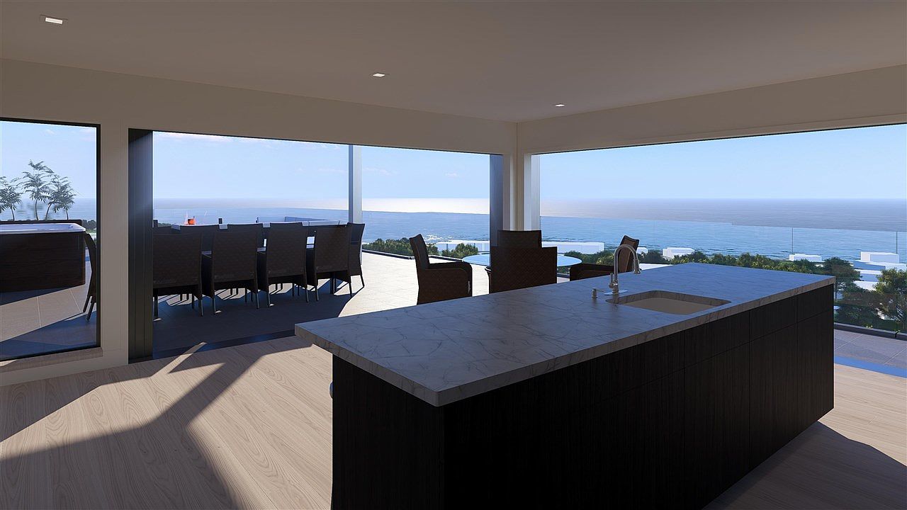 This is a bright and modern kitchen with a large island featuring a marble countertop and dark wood cabinetry. The kitchen opens to an outdoor dining area with stunning ocean views through large glass windows and sliding doors. The interior space is well-lit, creating an inviting and luxurious atmosphere.