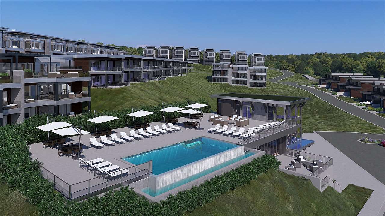 This image showcases a luxurious outdoor pool area with modern architectural elements. The pool features a waterfall edge and is surrounded by lounge chairs, tables with umbrellas, and lush greenery. The scene includes contemporary residential buildings in the background, suggesting a high-end community with resort-style amenities.