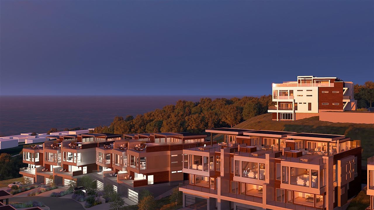 This aerial view showcases a modern residential complex with multiple units, each featuring private balconies and garages. The architecture emphasizes clean lines and large windows, offering stunning views of the ocean and surrounding landscape. The development is nestled on a hillside, blending seamlessly with the natural environment.