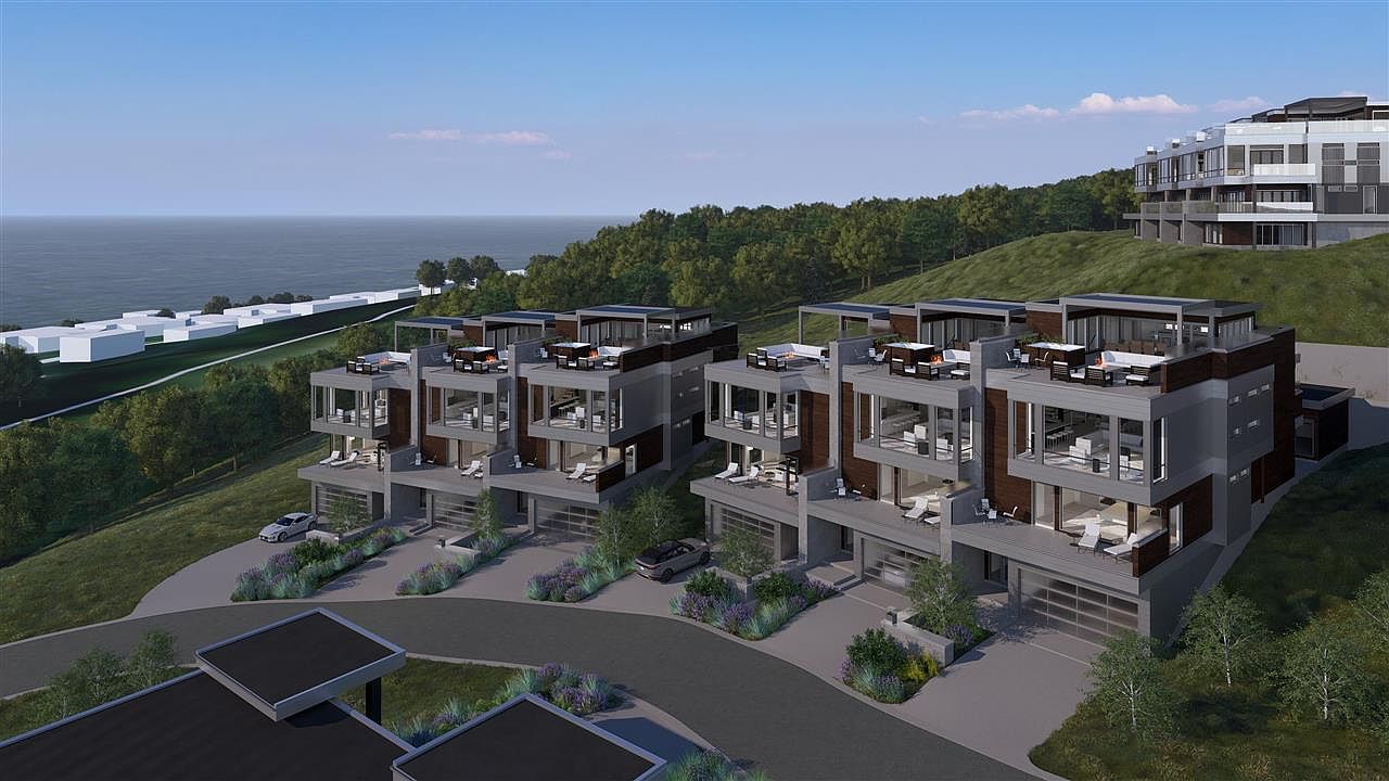 This aerial view showcases a set of modern townhouses nestled on a hillside overlooking the ocean. The townhouses feature a contemporary design with flat roofs, large windows, and private rooftop terraces furnished with outdoor seating. The surrounding landscape includes lush greenery and a winding driveway, creating an upscale and serene living environment.