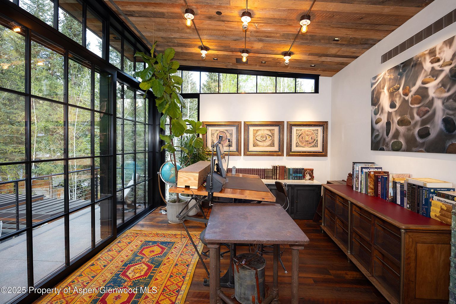 This interior shot showcases a well-appointed office space with a rustic yet sophisticated aesthetic. The room features a large window wall offering natural light and views of the surrounding greenery, complemented by a wooden plank ceiling with industrial-style lighting fixtures. The space is furnished with unique desks, a globe, and a built-in cabinet topped with books, creating a comfortable and inspiring work environment.