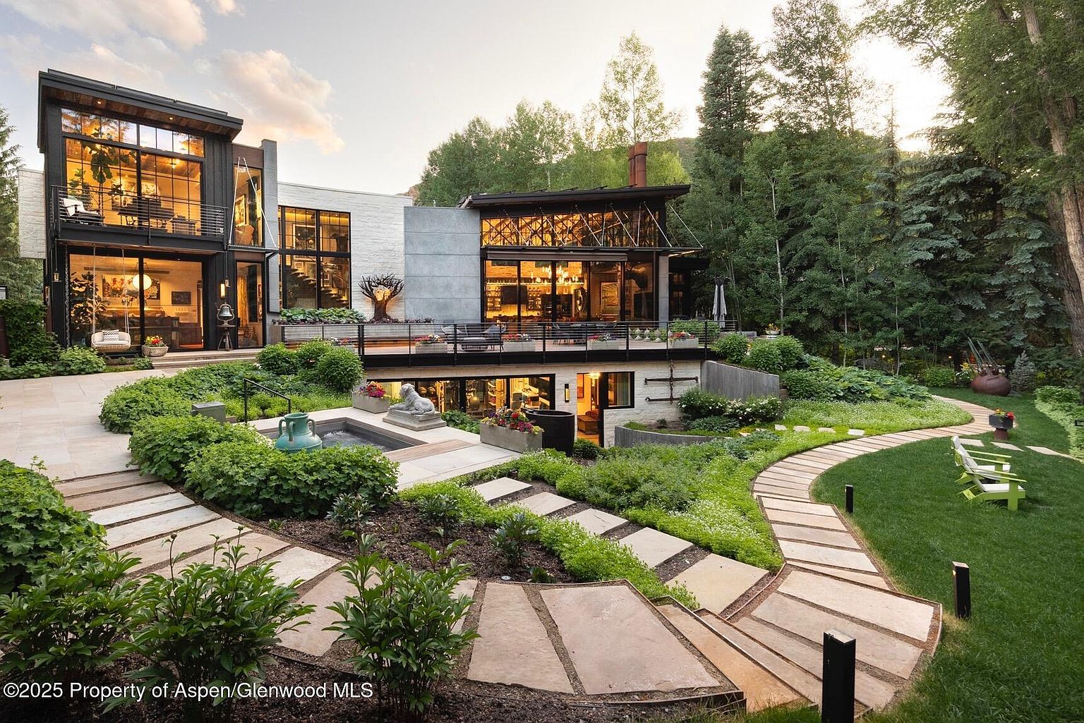 This image showcases the rear exterior of a modern home, emphasizing its integration with the surrounding landscape. The property features multiple levels with large windows, a stone pathway leading through lush gardens, and a patio area perfect for outdoor entertaining. The architecture blends contemporary design with natural elements, creating a serene and luxurious atmosphere.