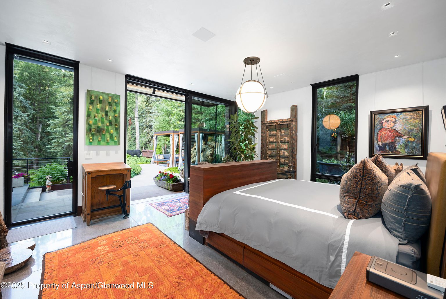 This is a primary bedroom featuring a modern platform bed with a gray duvet and decorative pillows. Large windows offer views of the lush outdoor greenery, enhancing the room's connection to nature. An orange rug adds a pop of color to the polished floor, and artwork adorns the walls, creating a sophisticated and tranquil atmosphere.