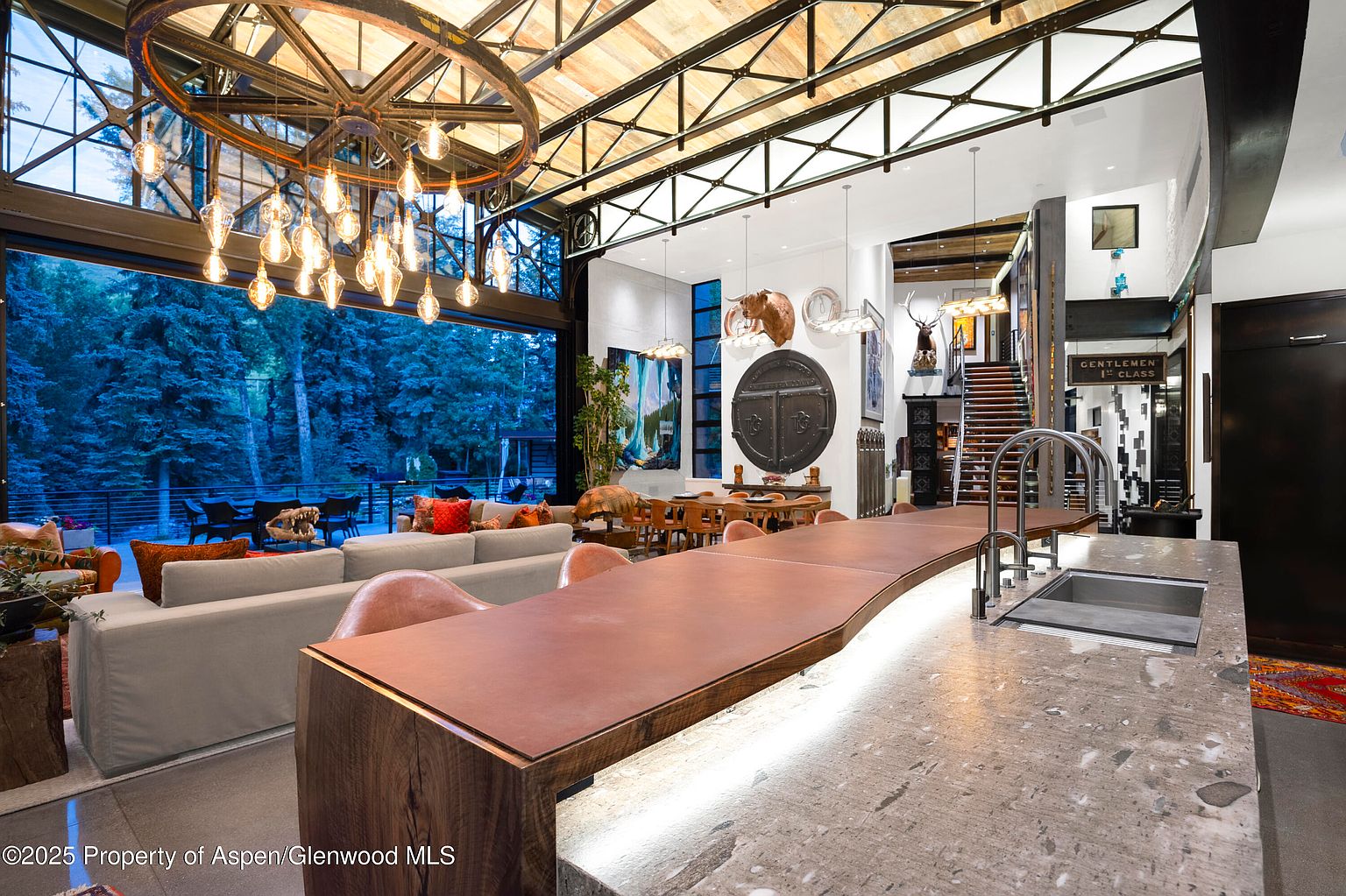 This interior shot showcases a luxurious living space with an open floor plan, seamlessly blending the living room, dining area, and kitchen. The room features a striking chandelier, a large sliding glass door that opens to an outdoor patio, and unique architectural details such as exposed beams and a custom bar. The overall impression is one of modern elegance and spaciousness, perfect for entertaining.