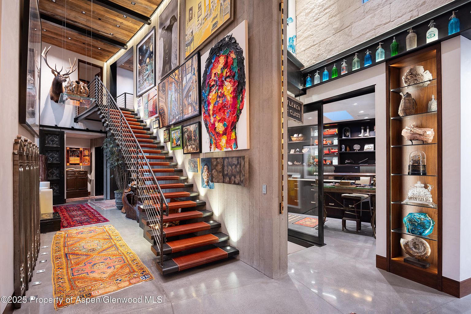 This interior shot showcases a grand hallway featuring a striking staircase with leather-clad steps and metal railings. The walls are adorned with an eclectic collection of artwork, adding a unique and artistic touch to the space. A display case filled with decorative objects and a glimpse into an office space further enhance the luxurious and sophisticated ambiance of the home.