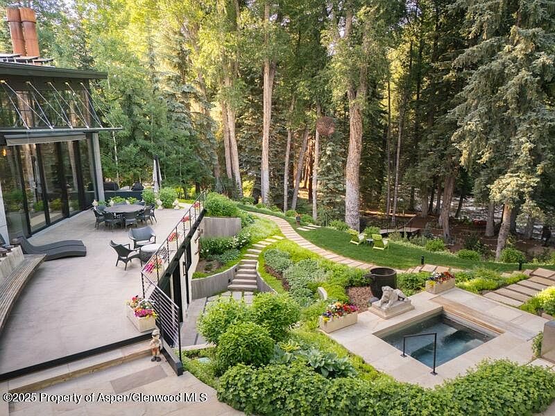 This image showcases a beautifully landscaped backyard featuring a modern home with large glass windows and a patio area. The yard includes a stone pathway, lush greenery, a hot tub, and mature trees, creating a serene and private outdoor space. The design blends contemporary architecture with natural elements, offering an inviting and luxurious atmosphere.