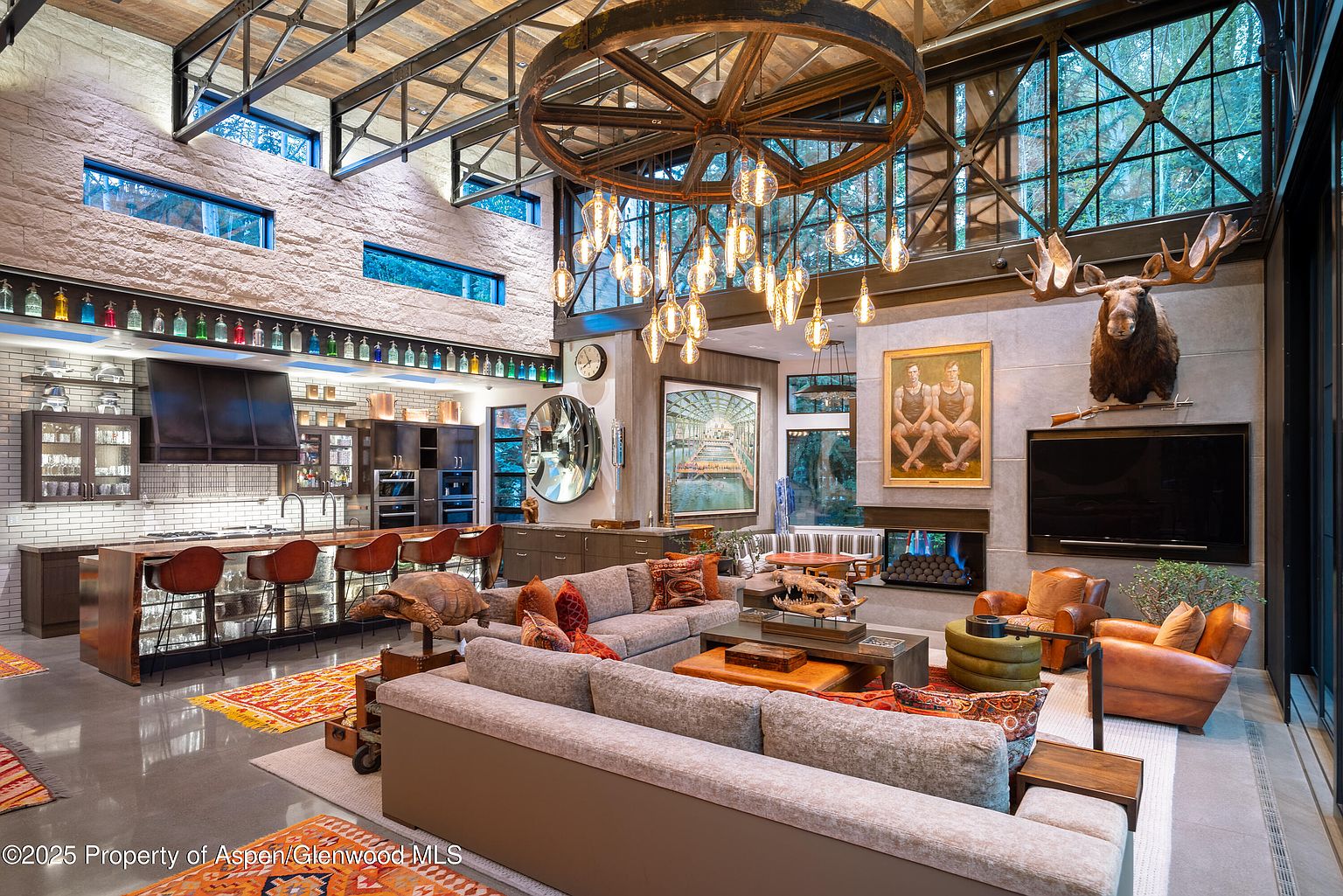 This expansive living room showcases a blend of rustic and modern design elements. A large, wheel-inspired chandelier hangs above a comfortable seating area with multiple sofas and armchairs. The room features a stone accent wall, a fireplace, and large windows offering ample natural light, creating a luxurious and inviting atmosphere.