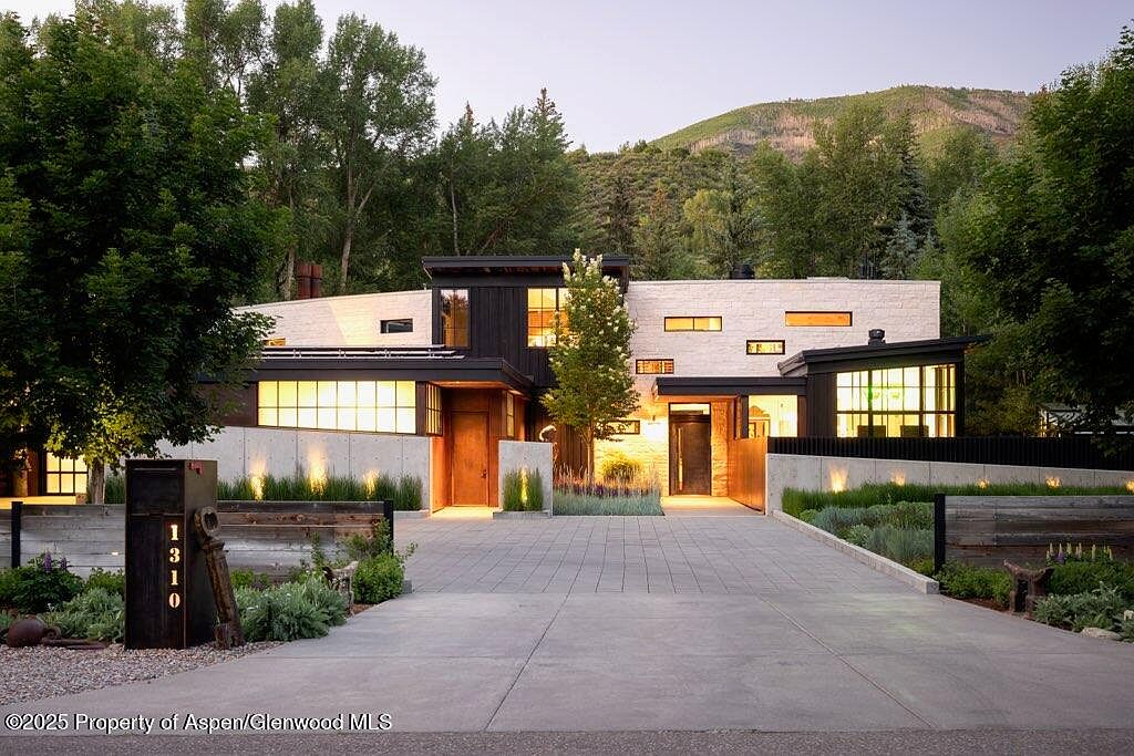This is a front view of a modern, luxurious home with a combination of light stone and dark wood accents. The property features a well-manicured landscape with a concrete driveway leading to the entrance. The architecture is contemporary, with clean lines and strategically placed windows, set against a backdrop of lush trees and a distant mountain.