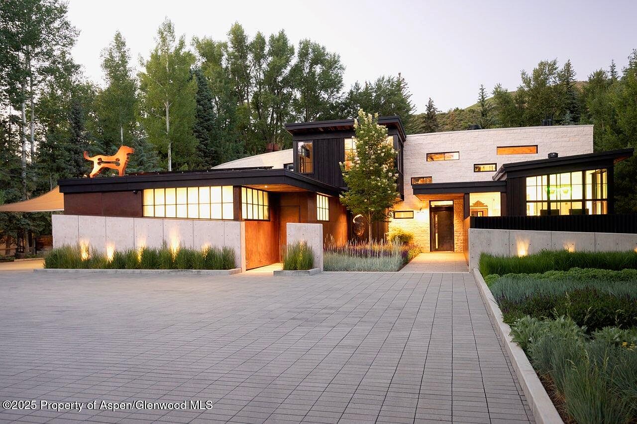 This is a front exterior view of a modern home with a unique architectural design. The property features a combination of concrete, wood, and dark metal elements, complemented by well-maintained landscaping. A paved driveway leads to the entrance, and the overall impression is one of contemporary elegance and curb appeal.